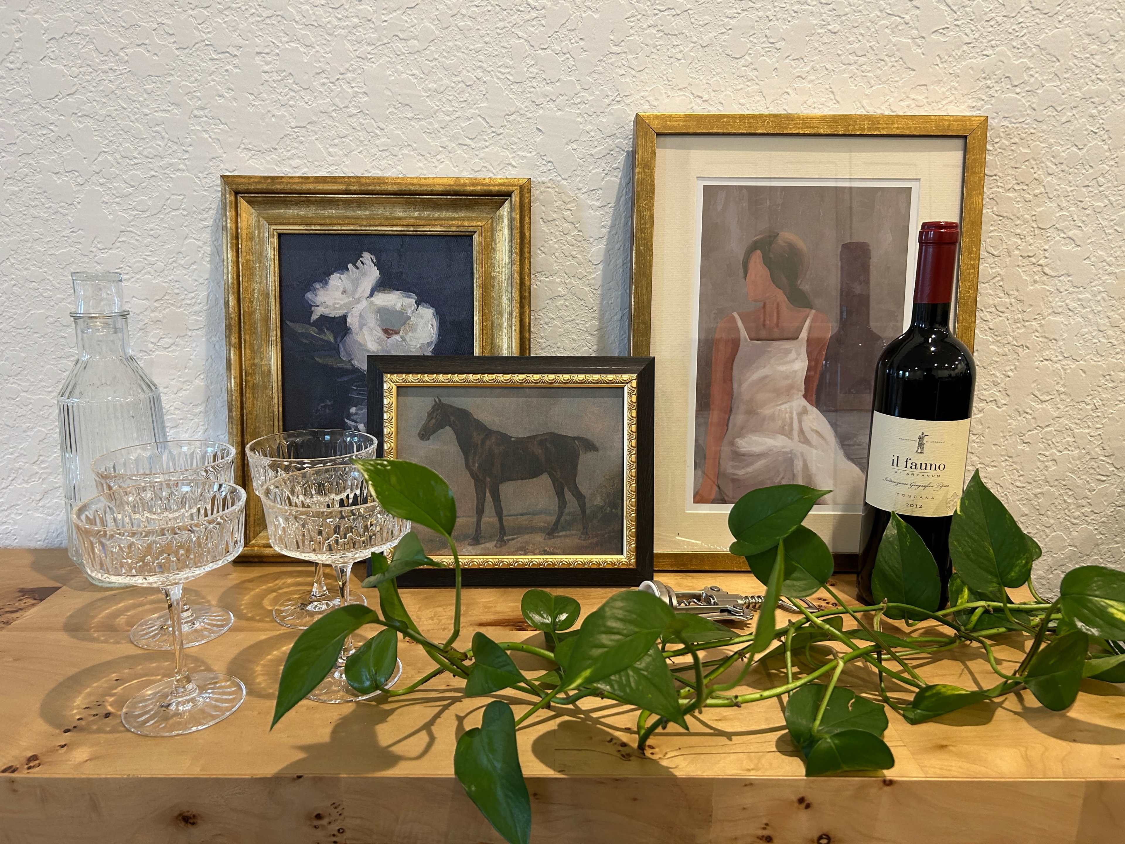 A wooden table displaying three framed artworks, two crystal glassware pieces, a bottle of wine, and a green vine.