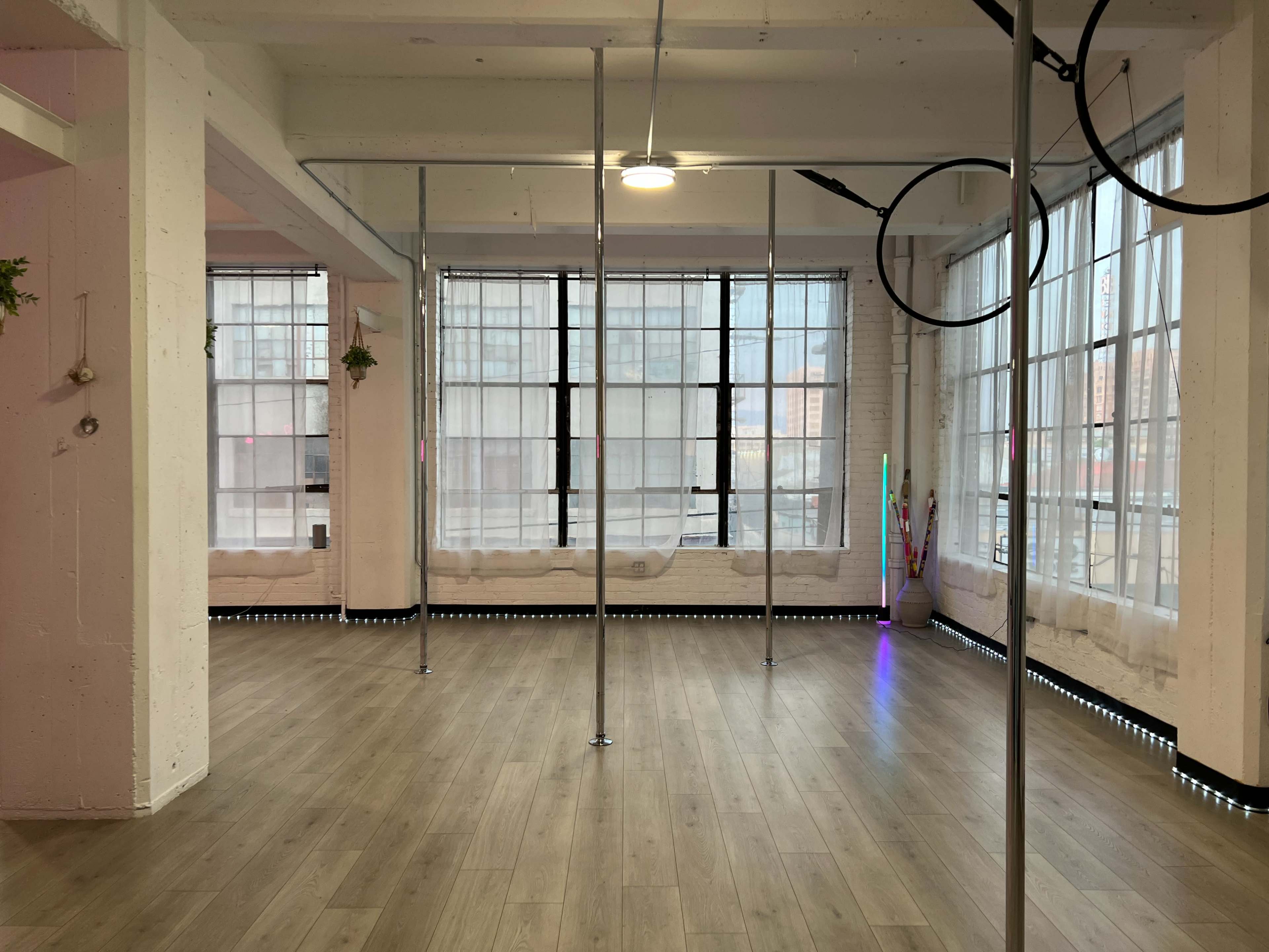The image shows a spacious interior with several vertical poles, large windows covered by sheer curtains, and wooden flooring.