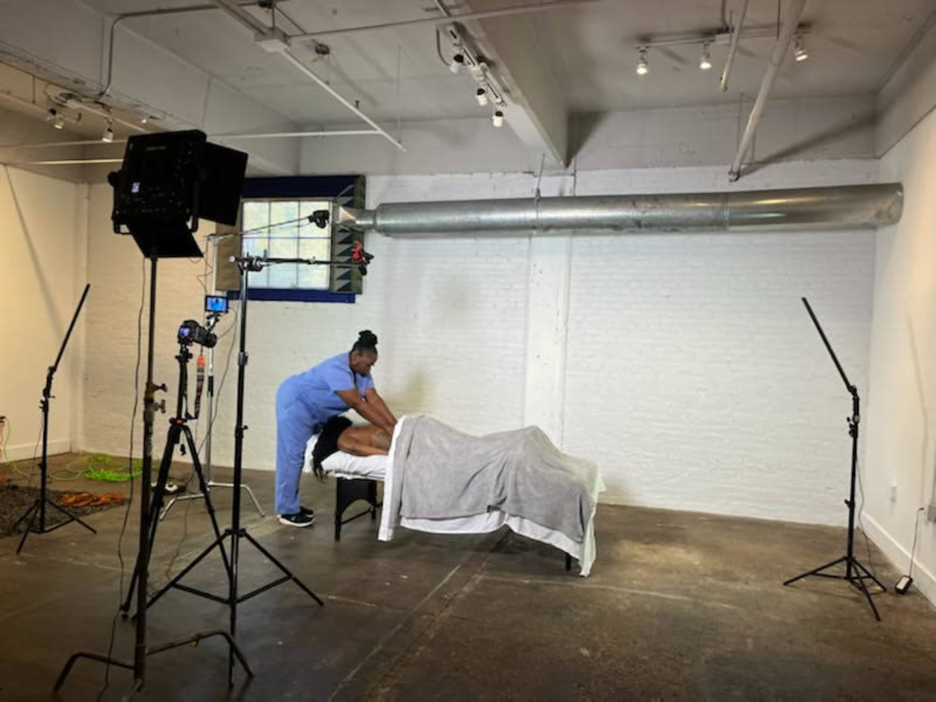 A person in scrubs is giving a massage to a client lying on a table in a well-lit room equipped with camera and lighting equipment.