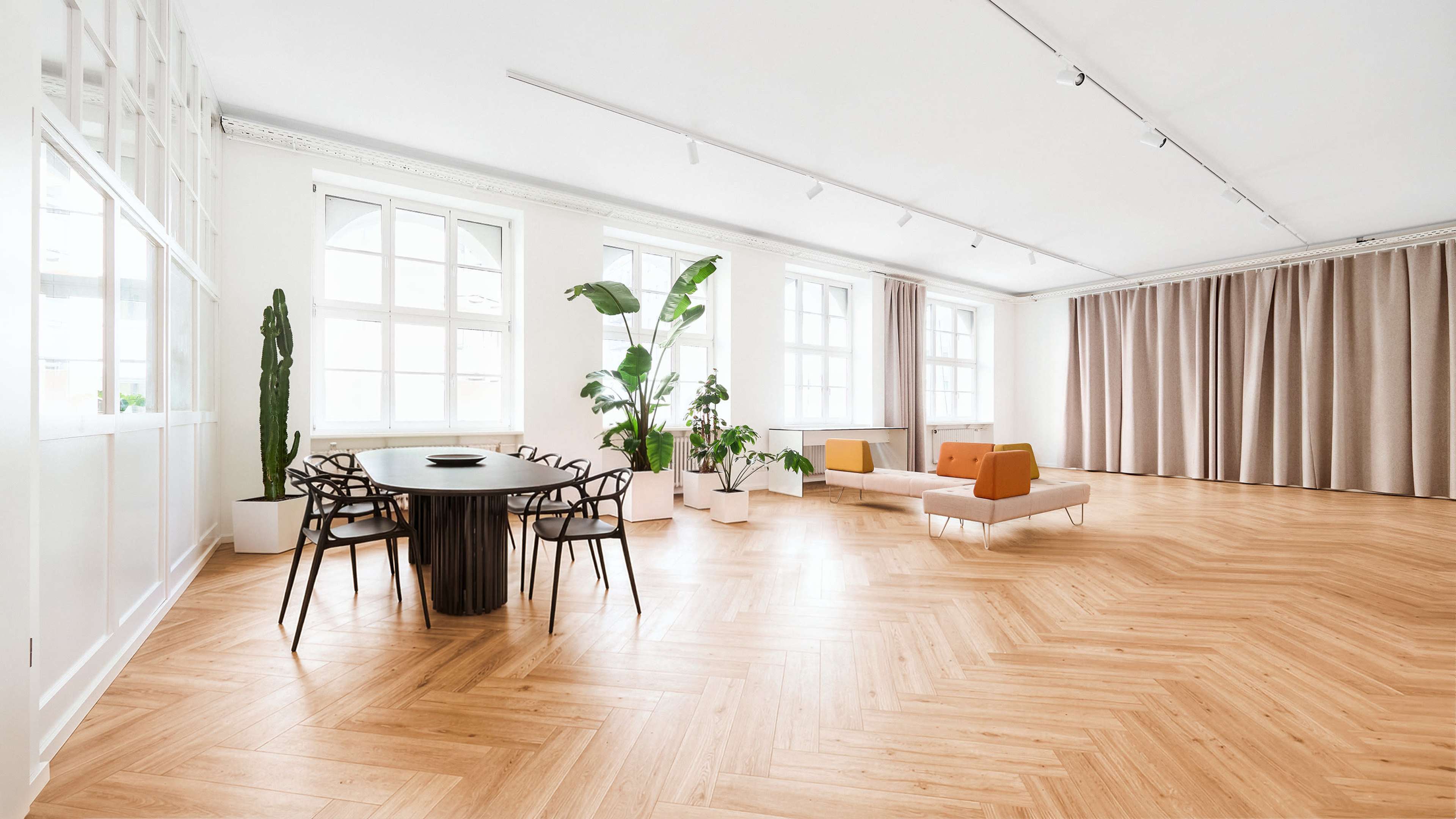 The image shows a spacious, modern interior with a wooden floor, a round black dining table with chairs, a large sofa in orange and beige, and several potted plants near large windows.