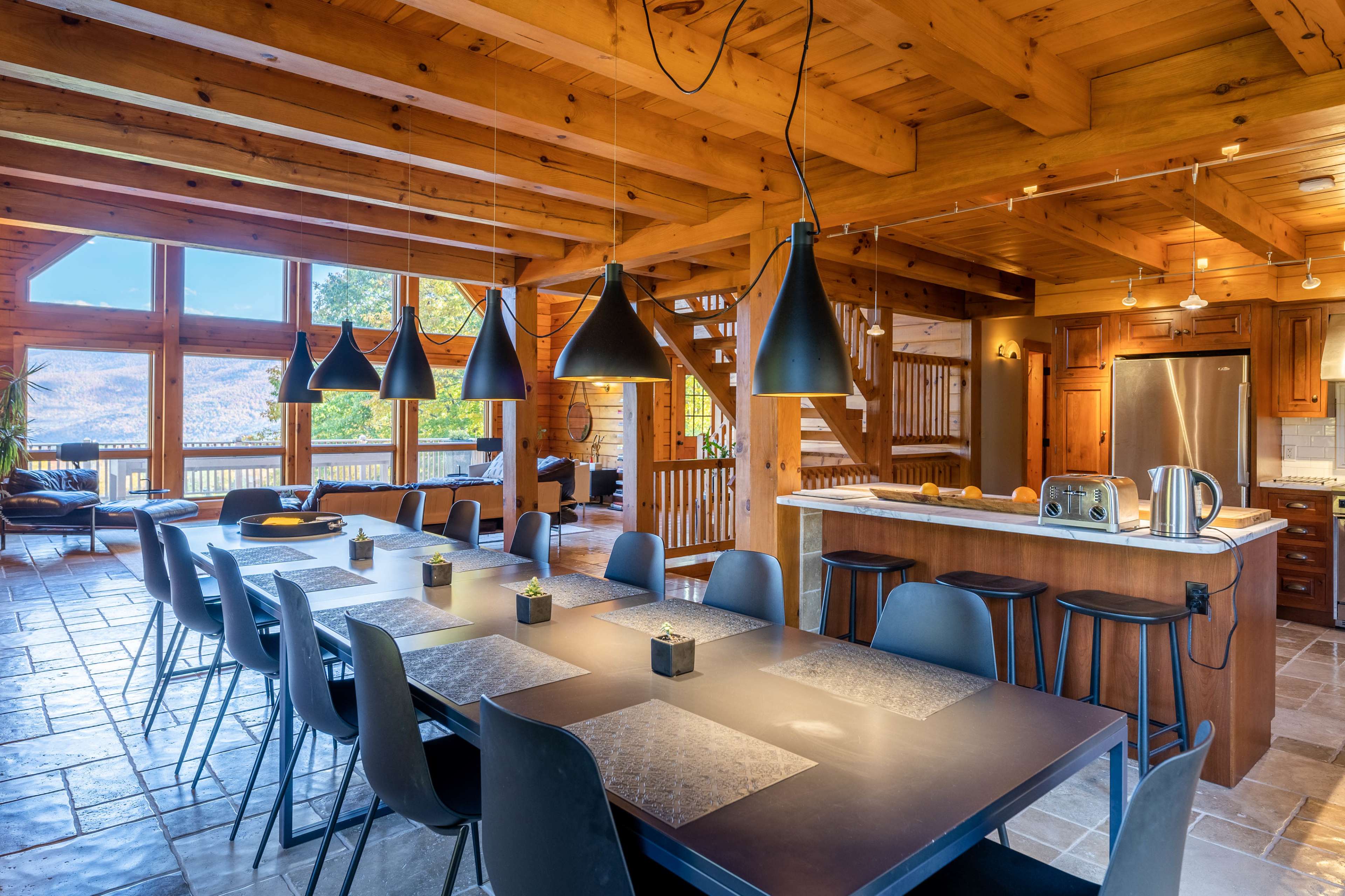 A spacious wooden dining area featuring a long table with black chairs, six hanging pendant lights, and a large window offering views of the outdoors.