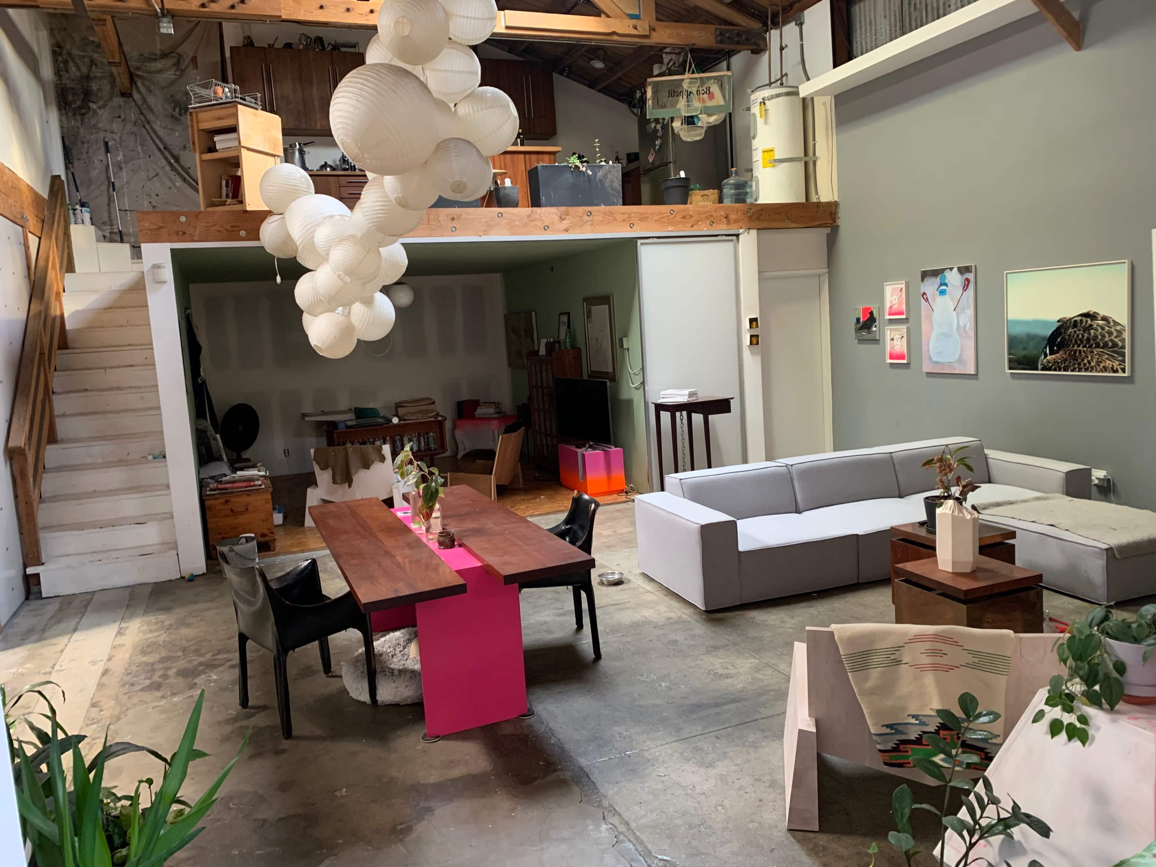 The interior of a spacious loft features a dining table with a pink runner, a gray sofa, and hanging paper lanterns above.
