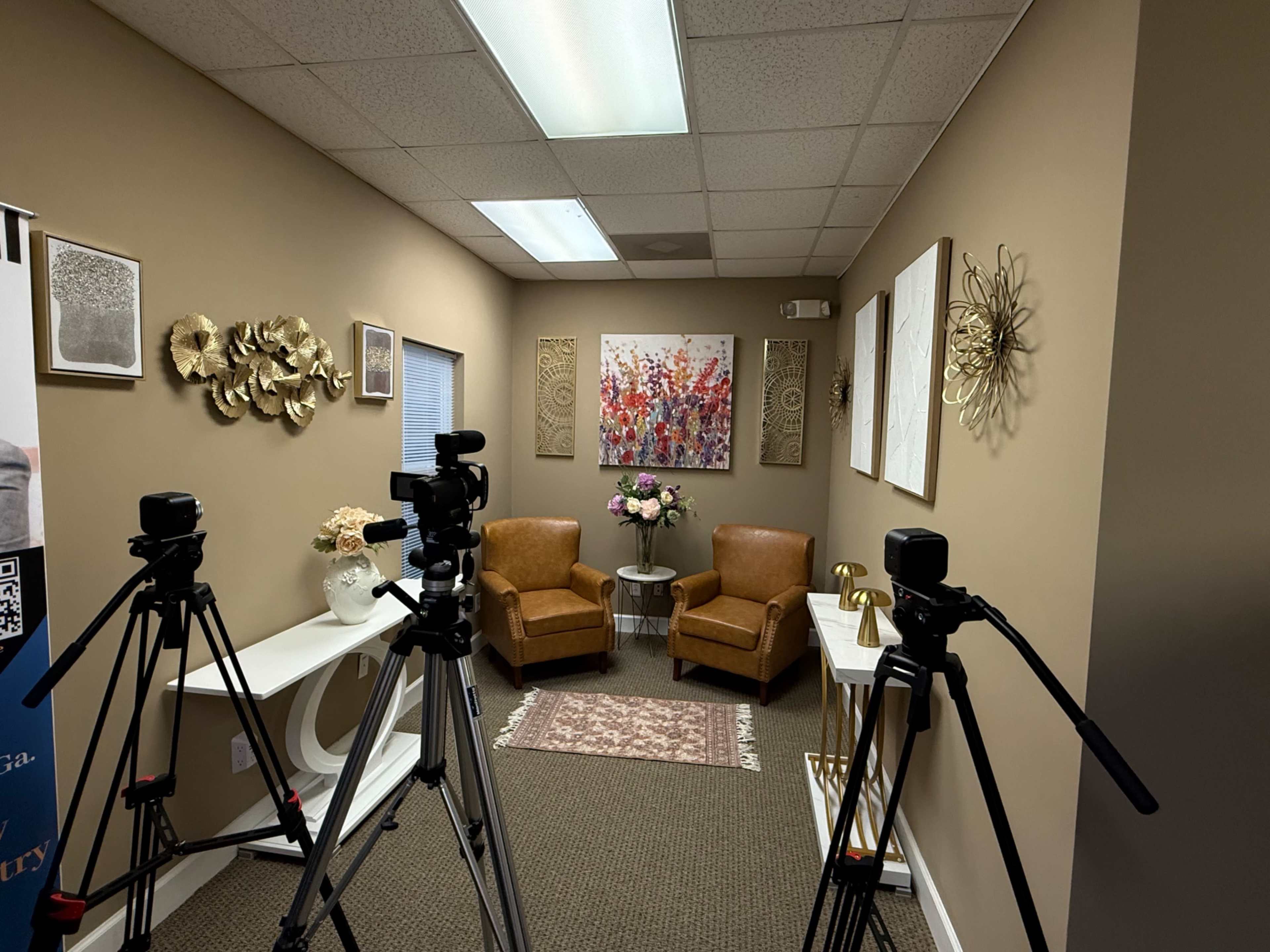 The image shows a small, professionally arranged room featuring two brown chairs, a decorative wall art piece, and cameras set up on tripods facing the seating area.