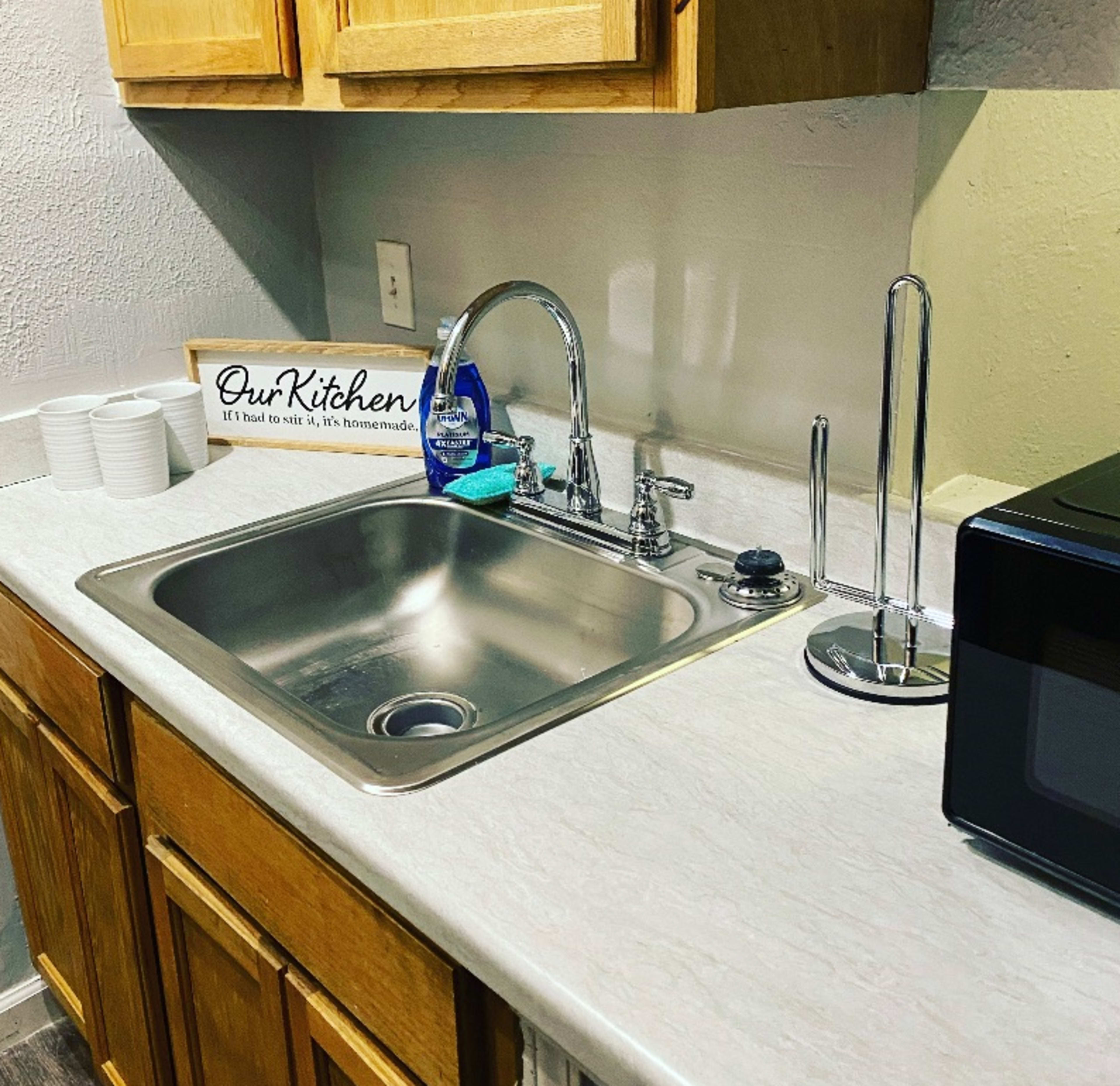 The image shows a kitchen sink with a silver faucet, a dish drying rack, a microwave, and wooden cabinets above a light-colored countertop.