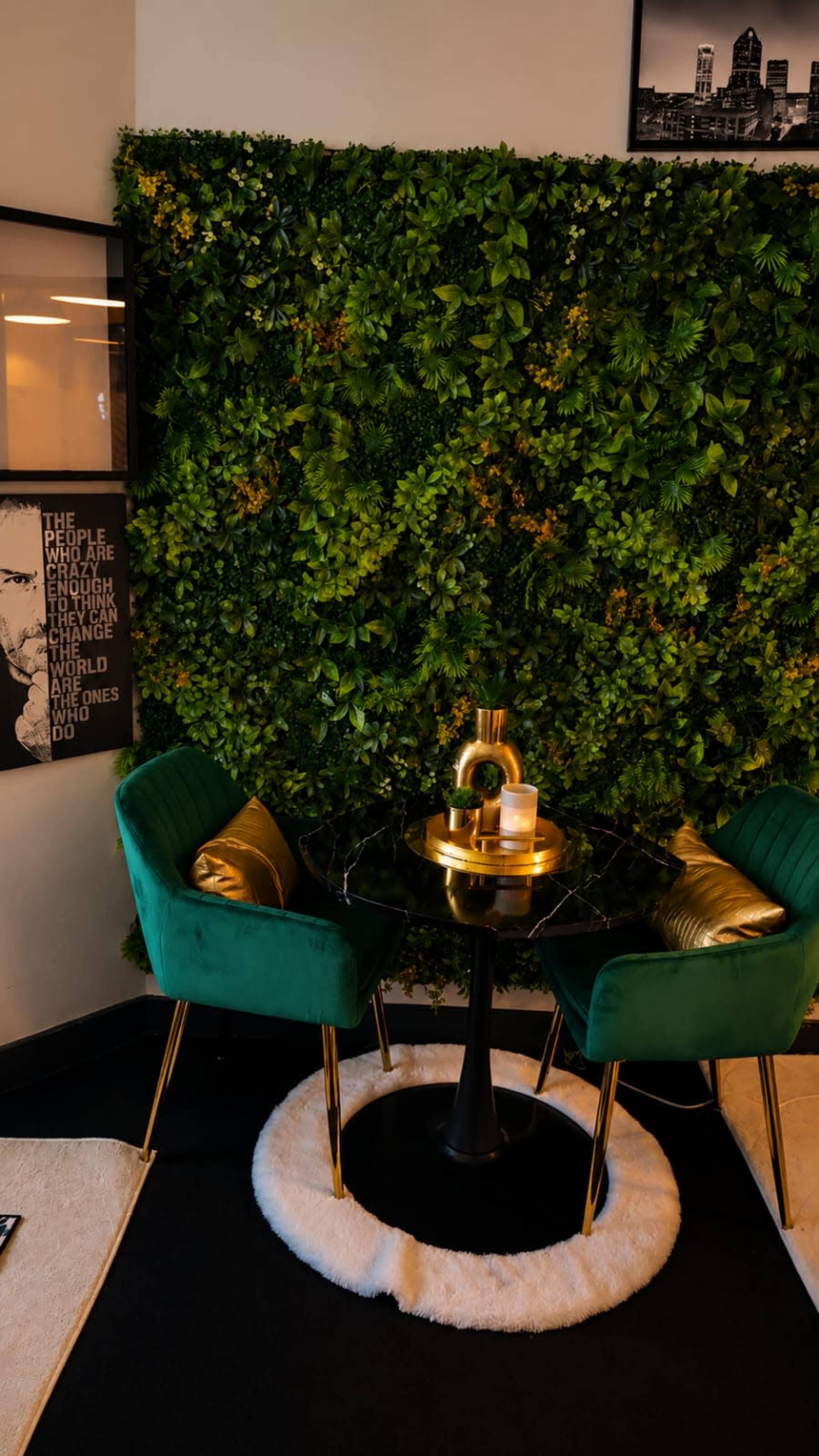 A cozy seating area features two green velvet chairs beside a black round table, set against a vibrant green wall of foliage.