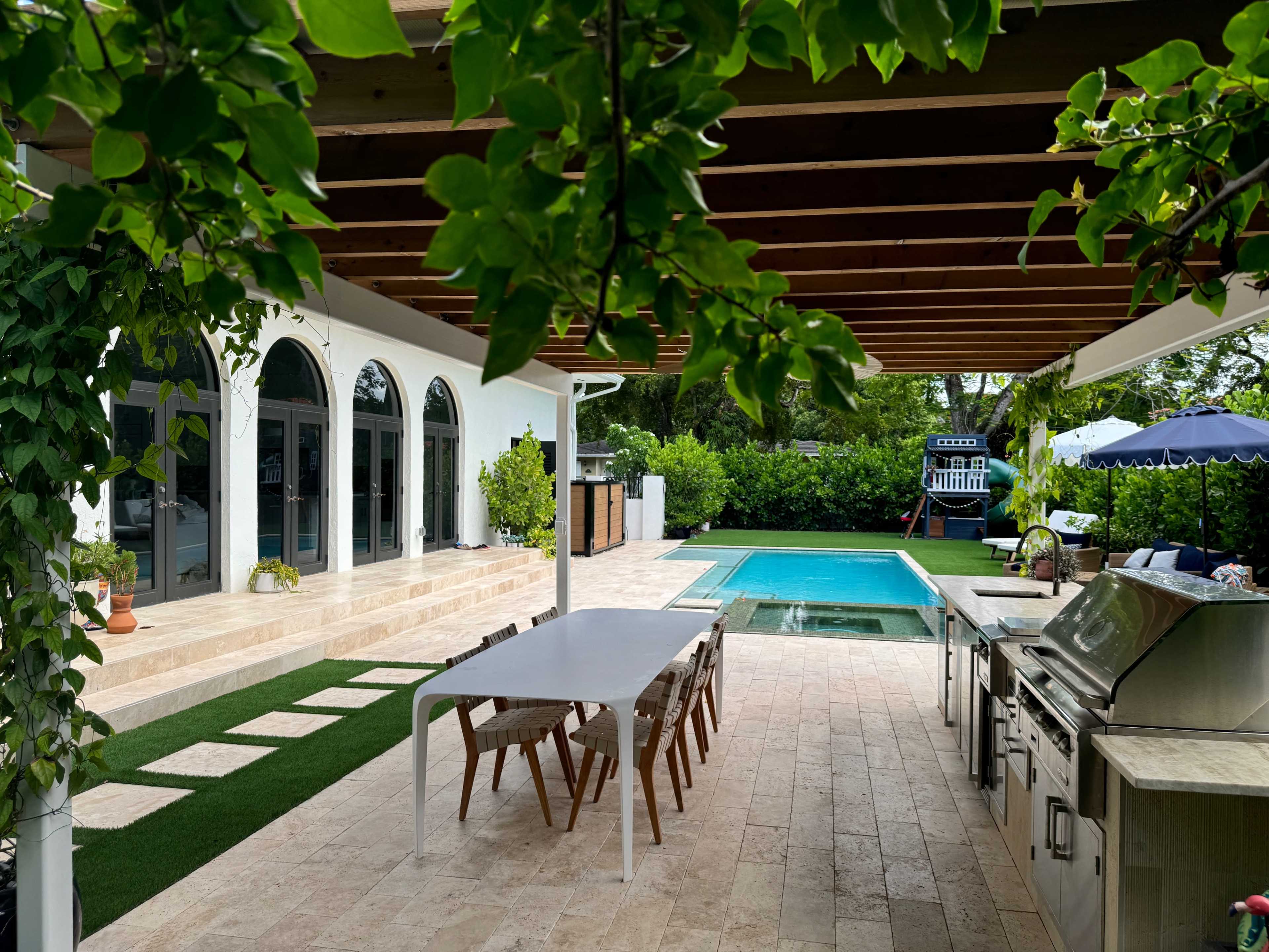 The image shows a backyard with a pool, outdoor kitchen, and seating area under a wooden pergola, surrounded by greenery.