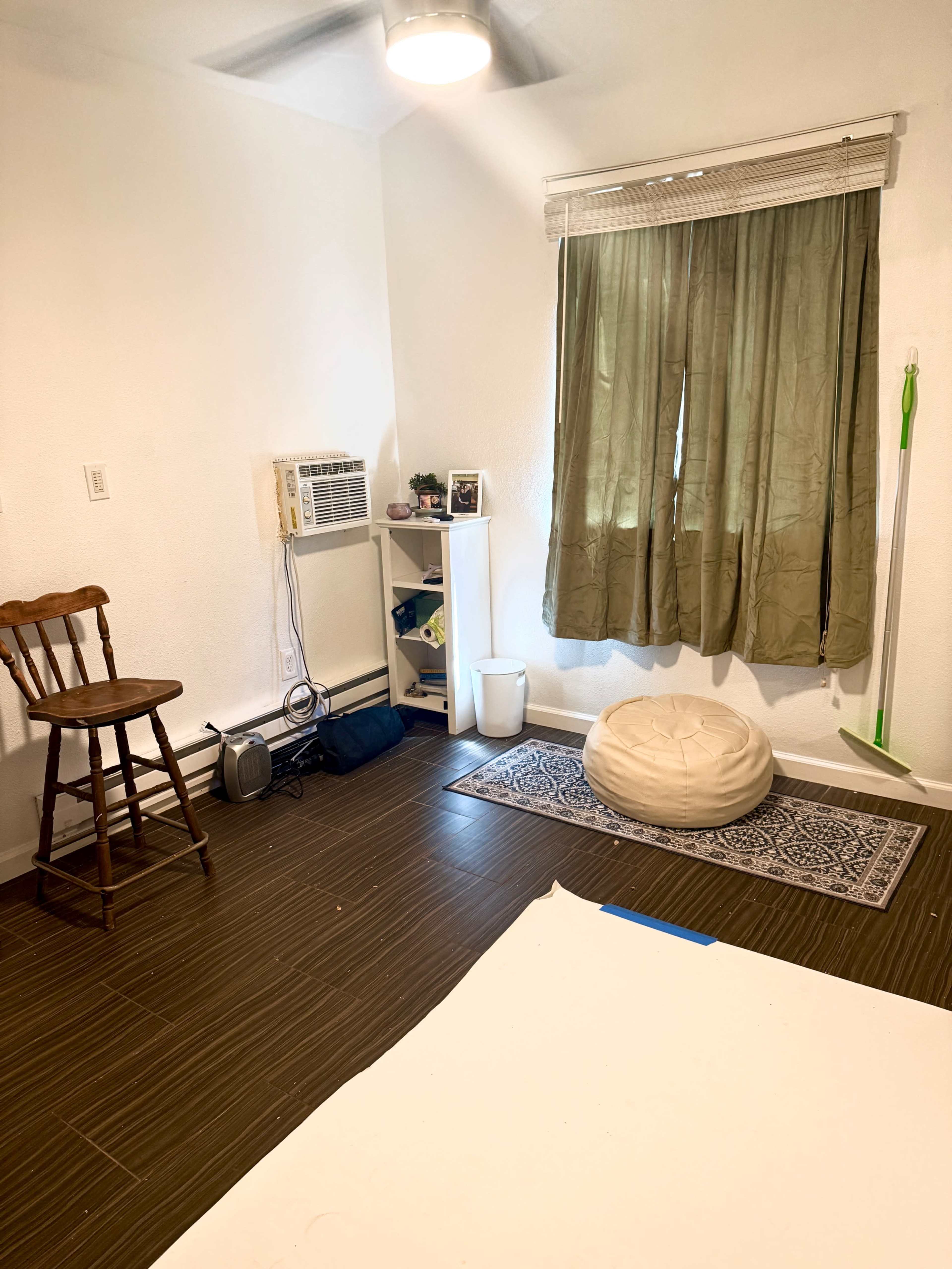 The image shows a small, sparsely furnished room with a chair, an air conditioning unit, a shelf, a round pouf, and a window covered by green curtains.