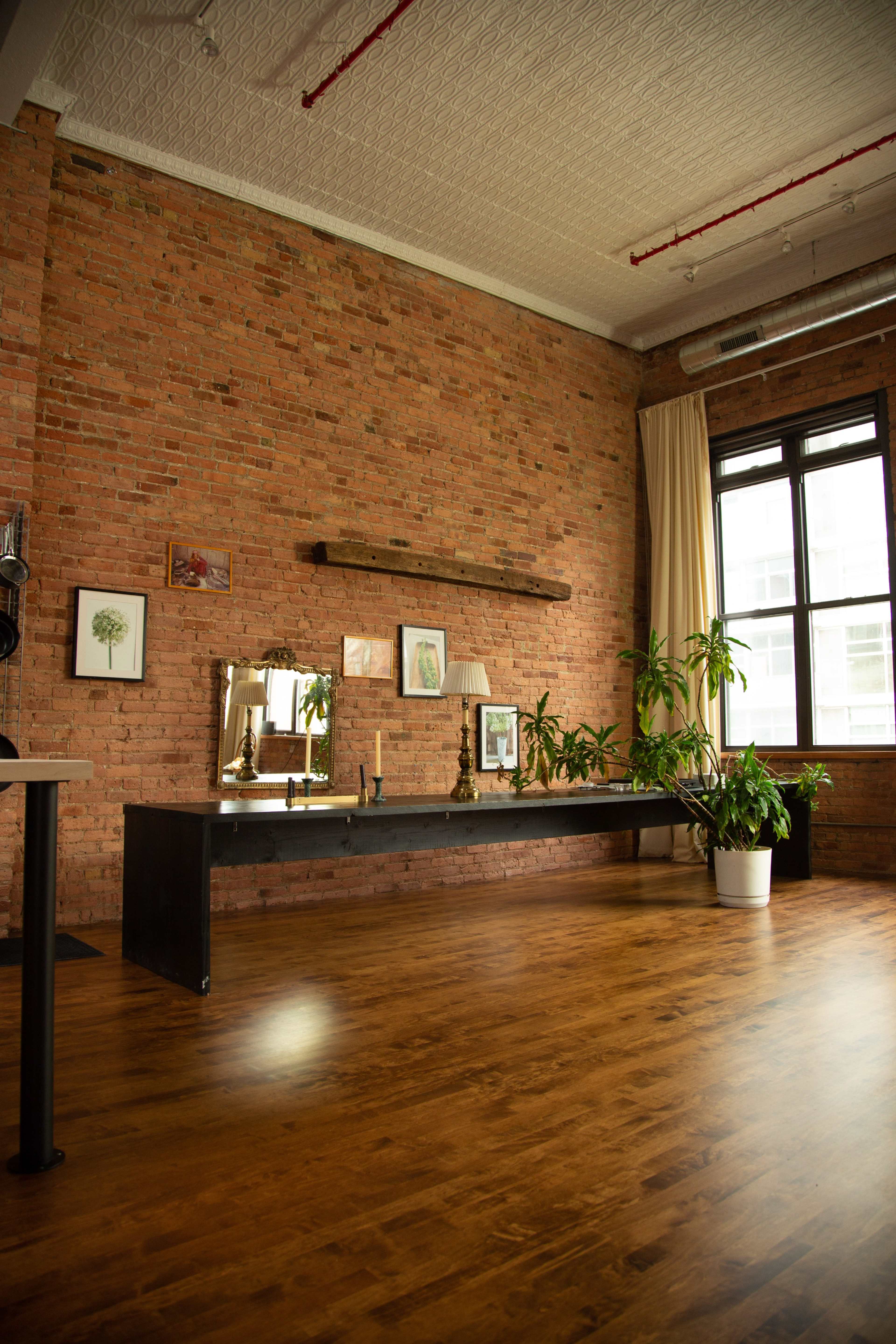 The image shows a spacious room with a brick wall, a long wooden table, a mirror, and a potted plant near a large window.