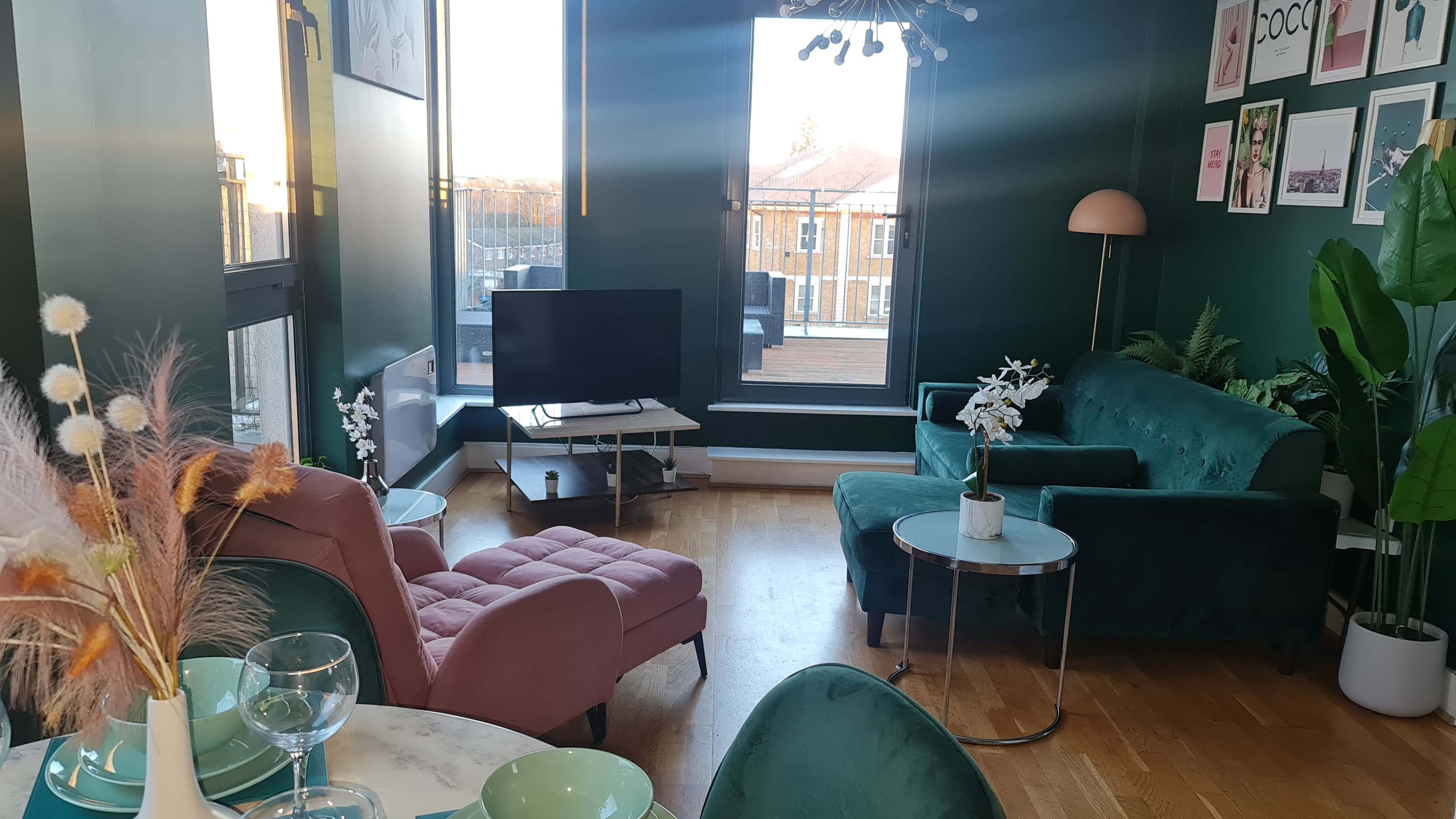 The scene features a modern living room with green walls, a pink armchair, a green sofa, a small round table, and a television set, all illuminated by natural light coming from large windows.