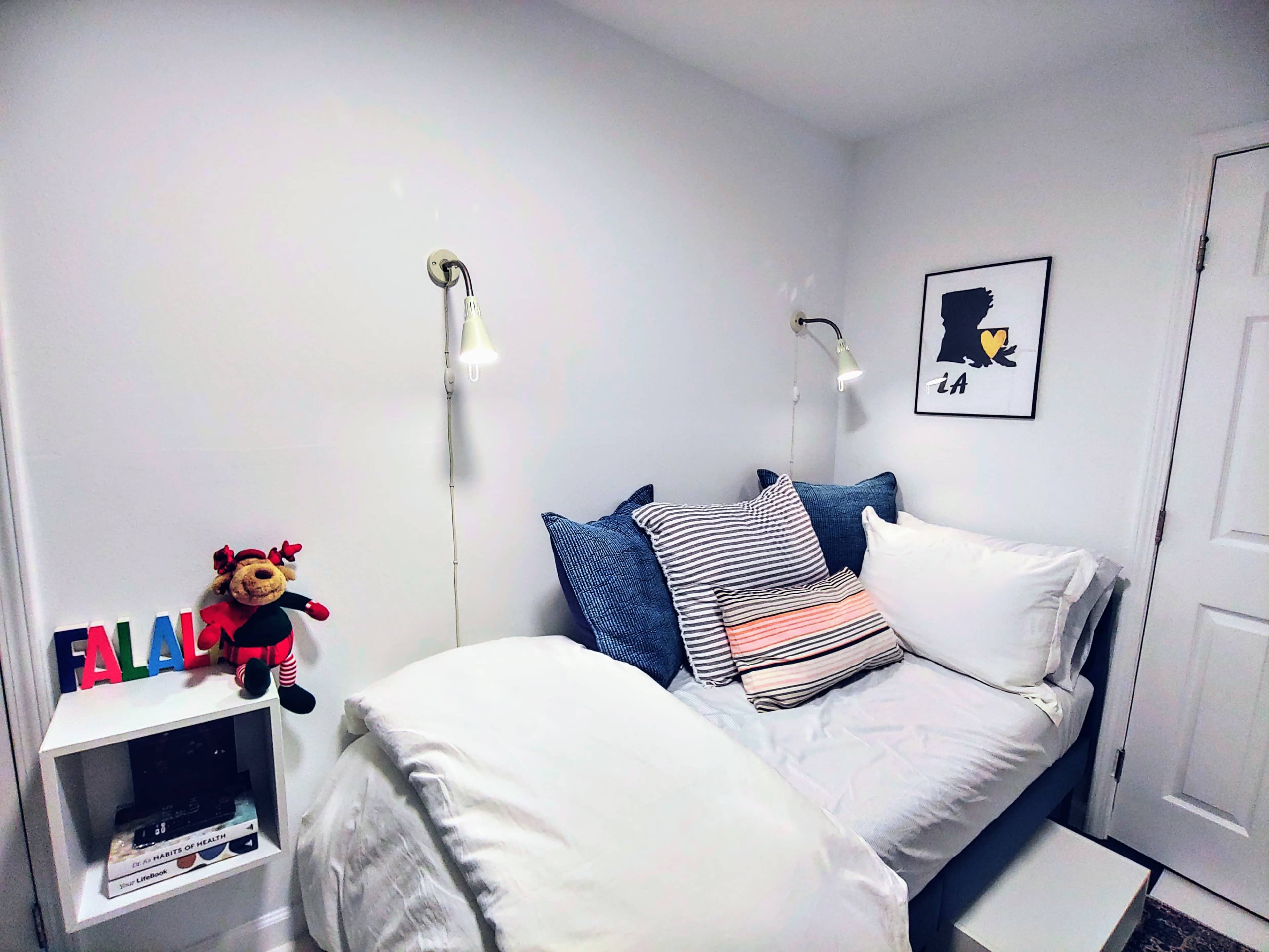 The image shows a neatly arranged bedroom with a single bed, decorative pillows, a framed artwork on the wall, and a nightstand featuring colorful letters and a stuffed toy.