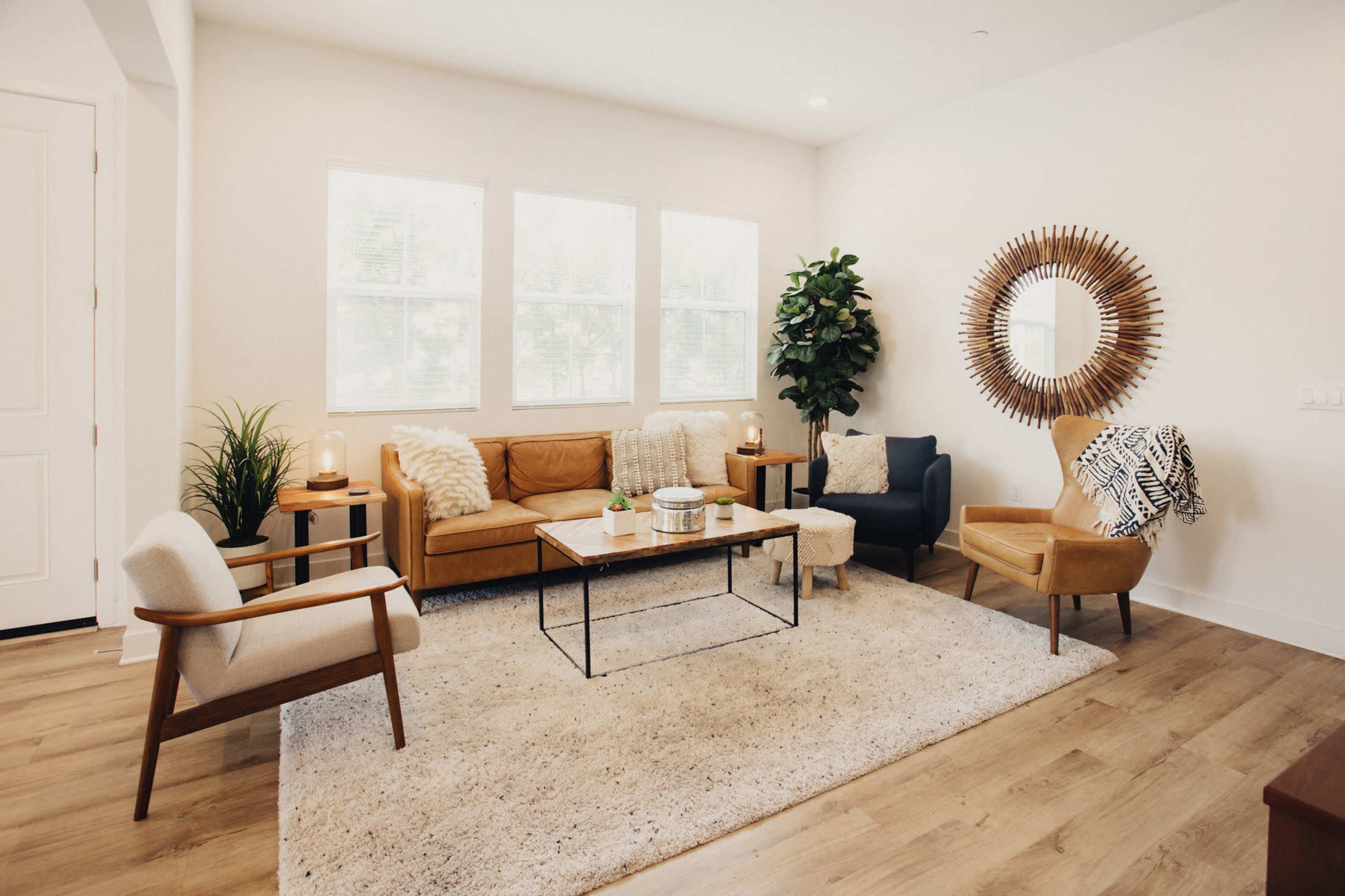 A modern living room furnished with a brown leather sofa, two accent chairs, a coffee table, and a large decorative mirror, all arranged on a light-colored rug.