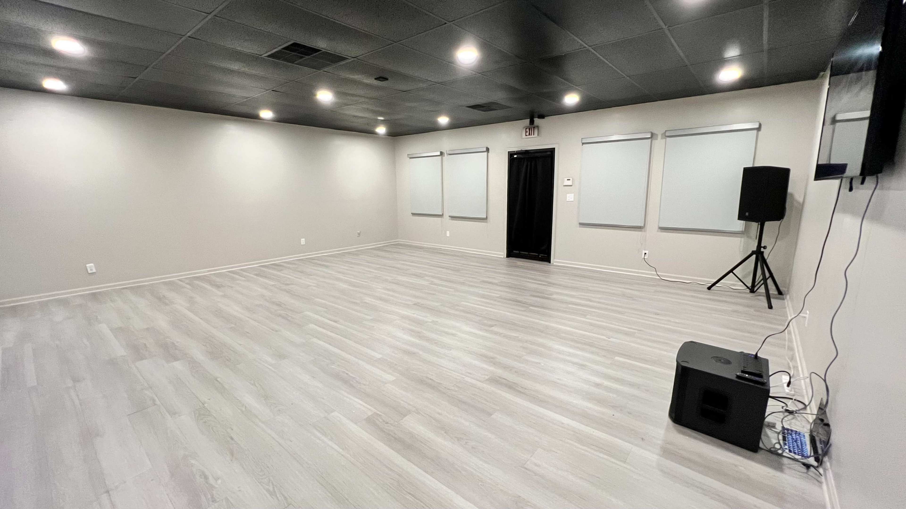 The image shows a spacious room with gray walls and flooring, equipped with a black speaker, a dark door, and several windows covered by light-colored shades.
