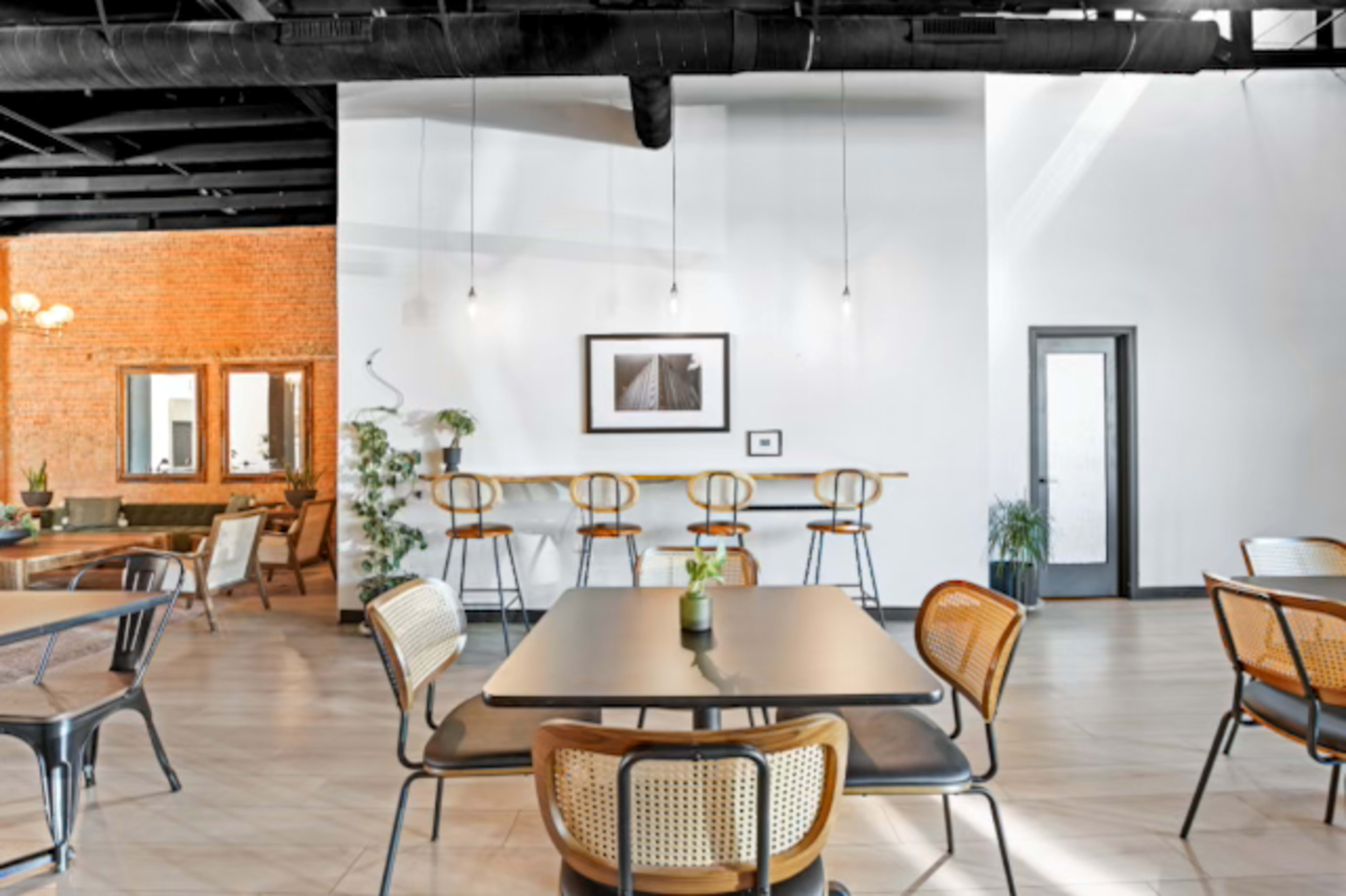 The image shows a spacious, modern dining area with minimalist decor, featuring a central table surrounded by chairs and a bar with stools in the background.