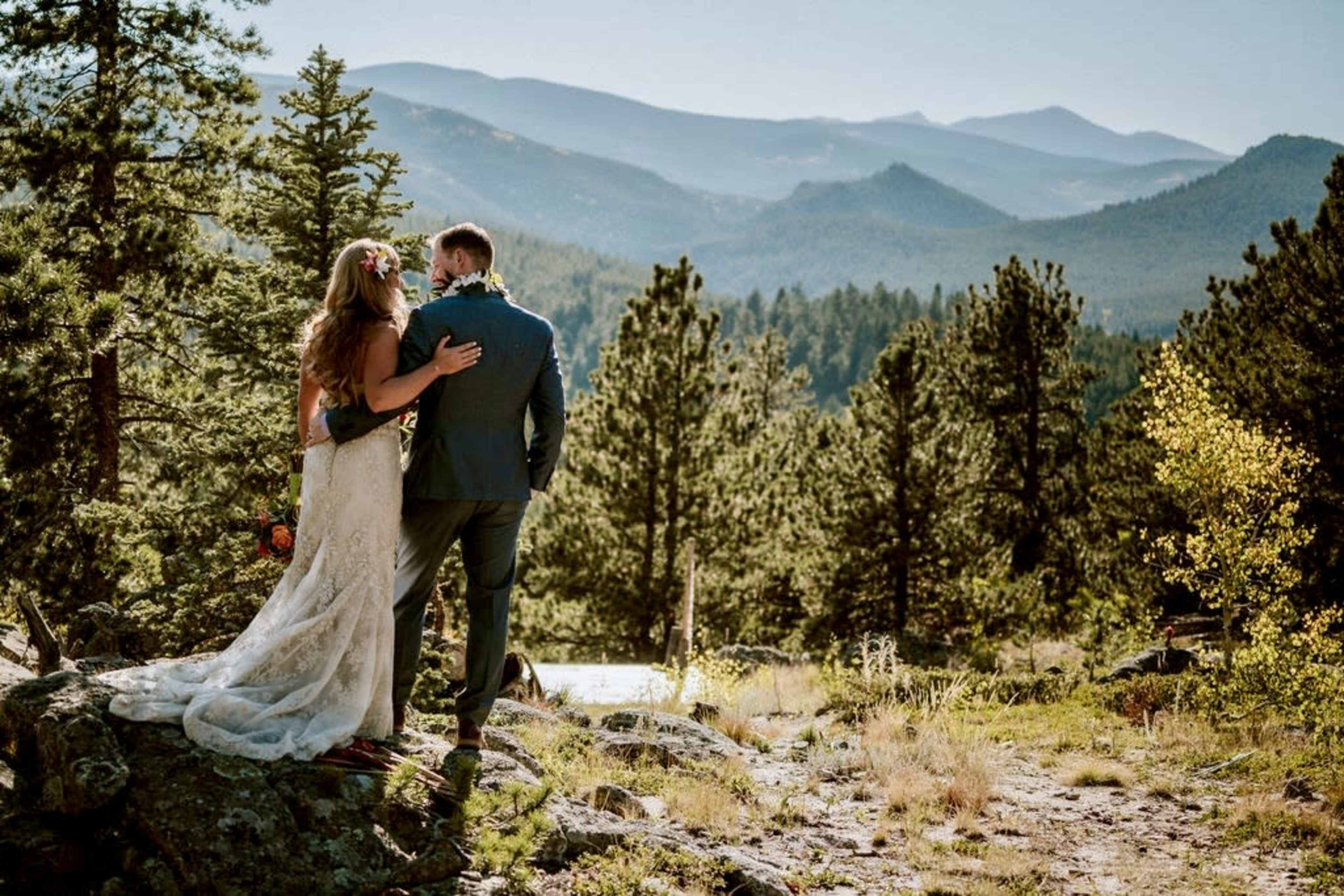 A bride and groom stand together on a rocky outcrop, overlooking a mountainous landscape surrounded by trees.