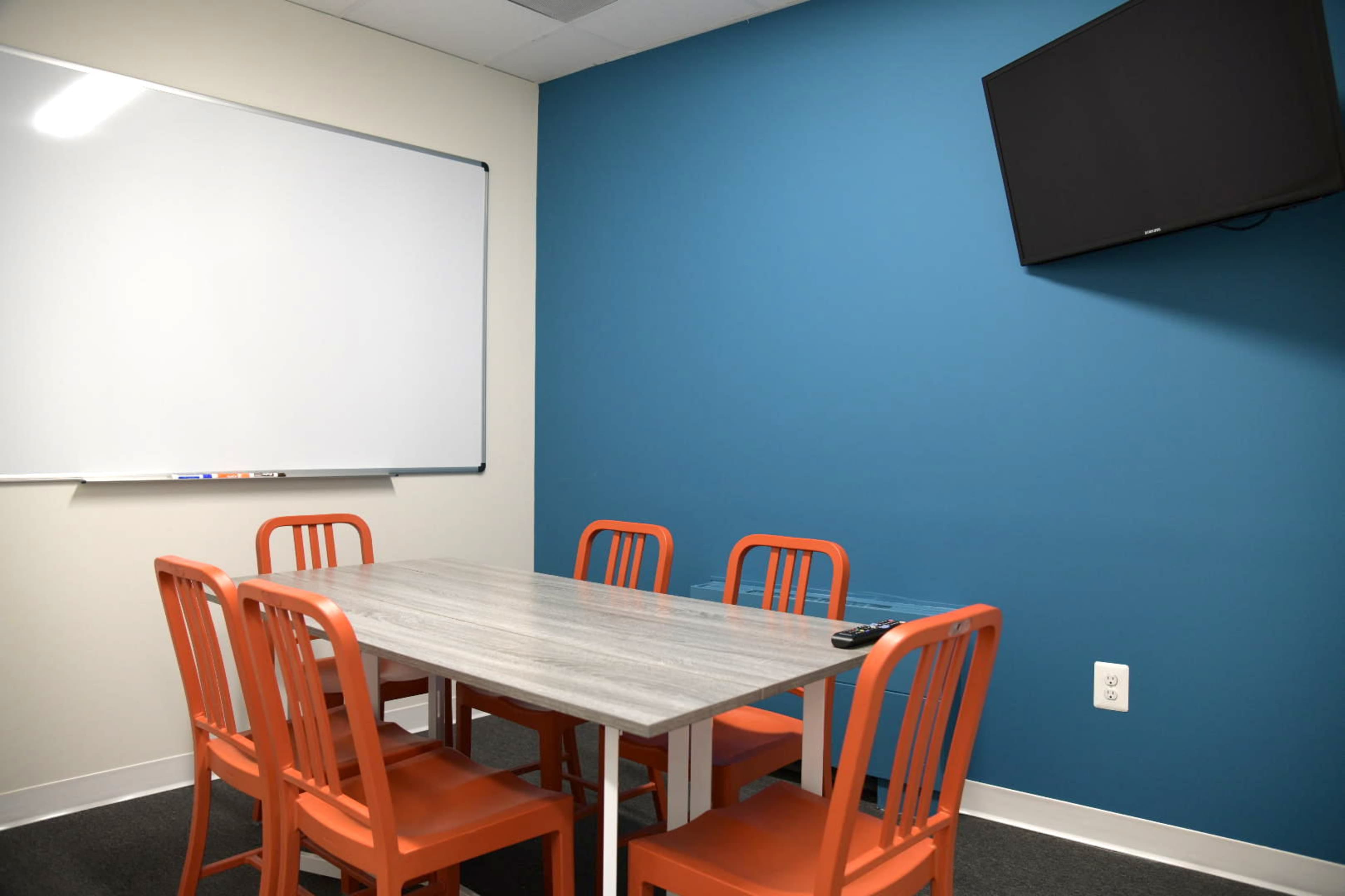 A small meeting room with a gray table, six orange chairs, a whiteboard on one wall, and a mounted television on another wall.