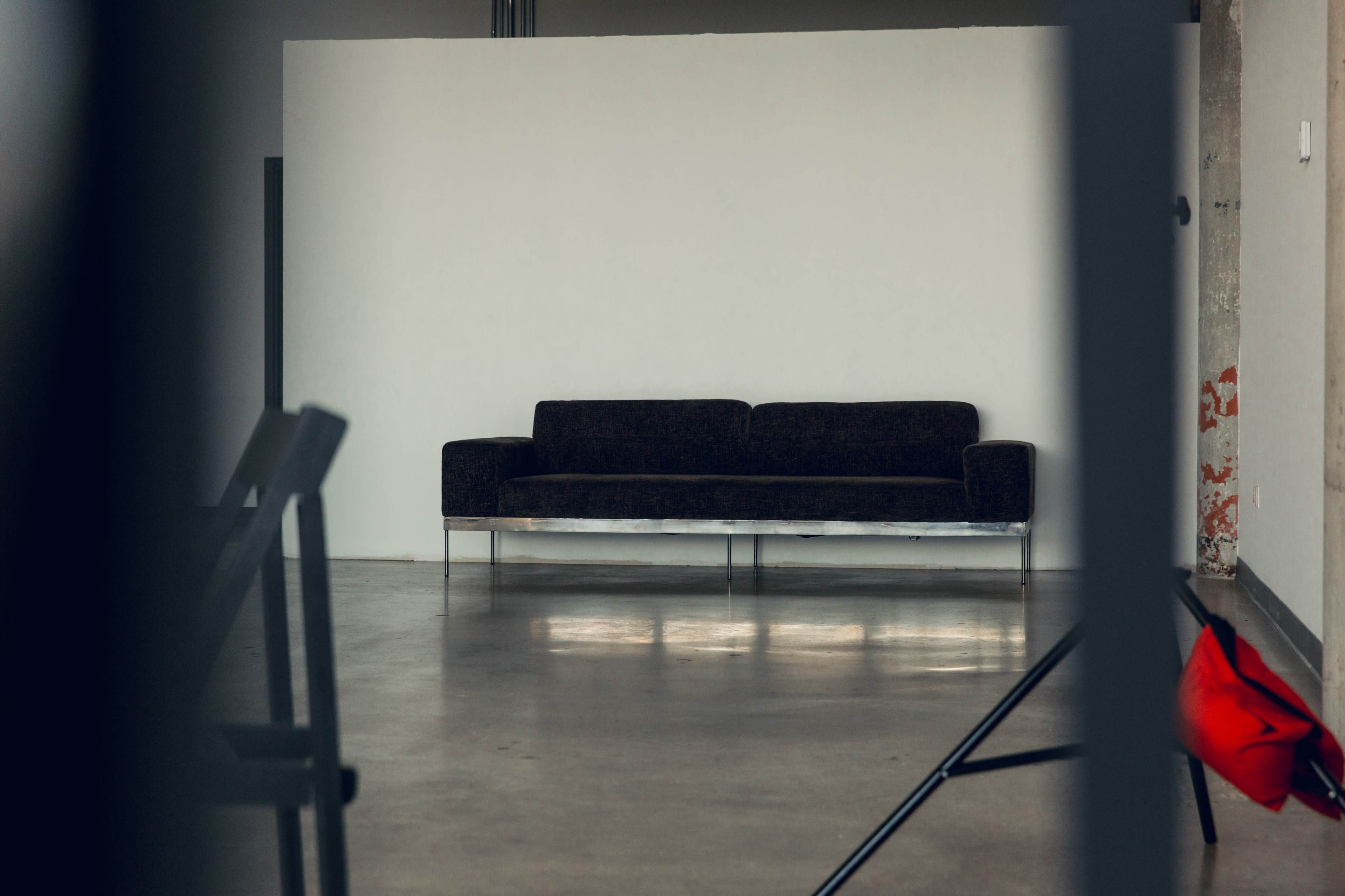 A dark-colored sofa against a white wall in an empty room with concrete flooring.