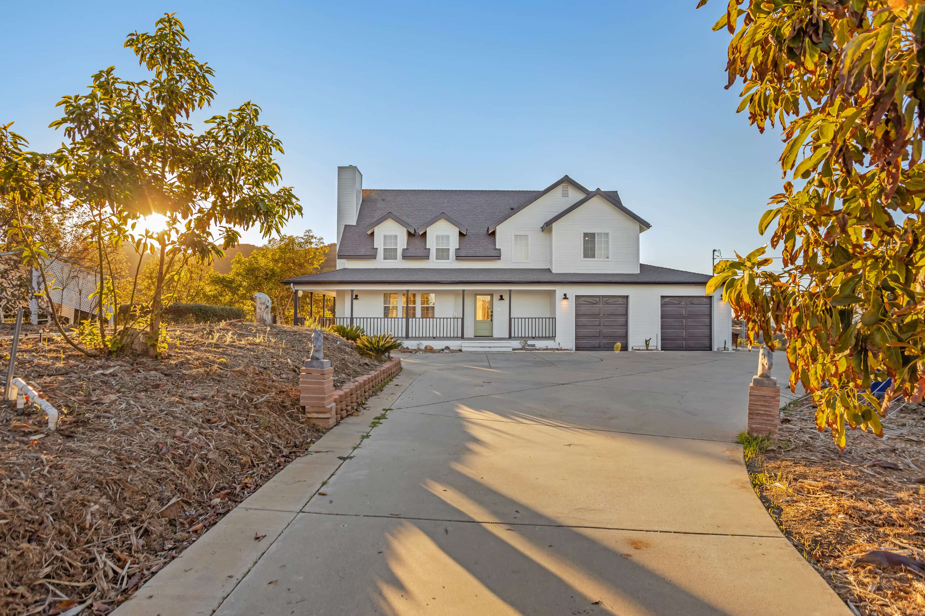 A two-story white house with a dark roof and a driveway is surrounded by greenery and sunlight.