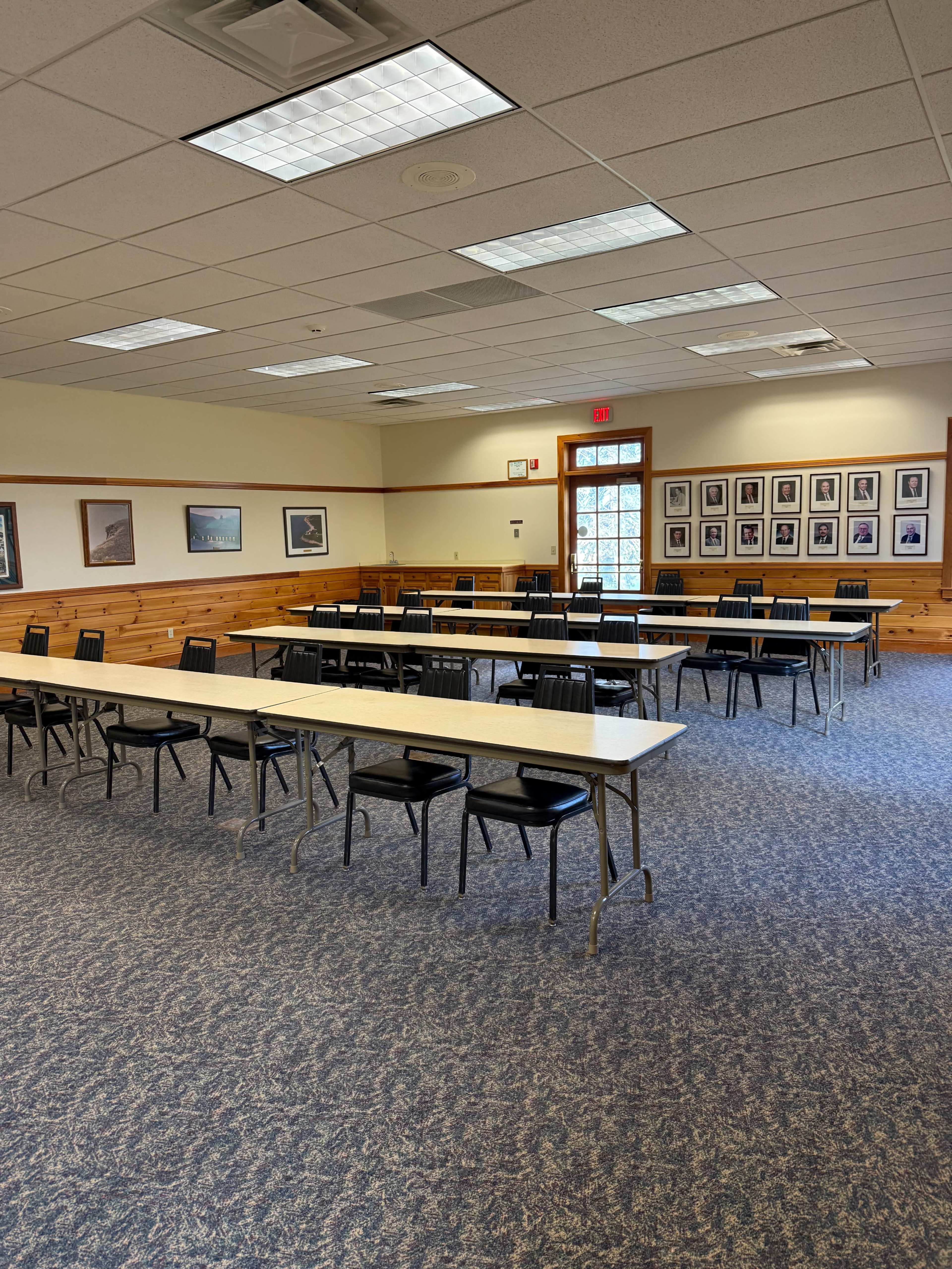 The image shows a spacious room arranged with several long tables and black chairs, featuring wood-paneled walls and framed photos.