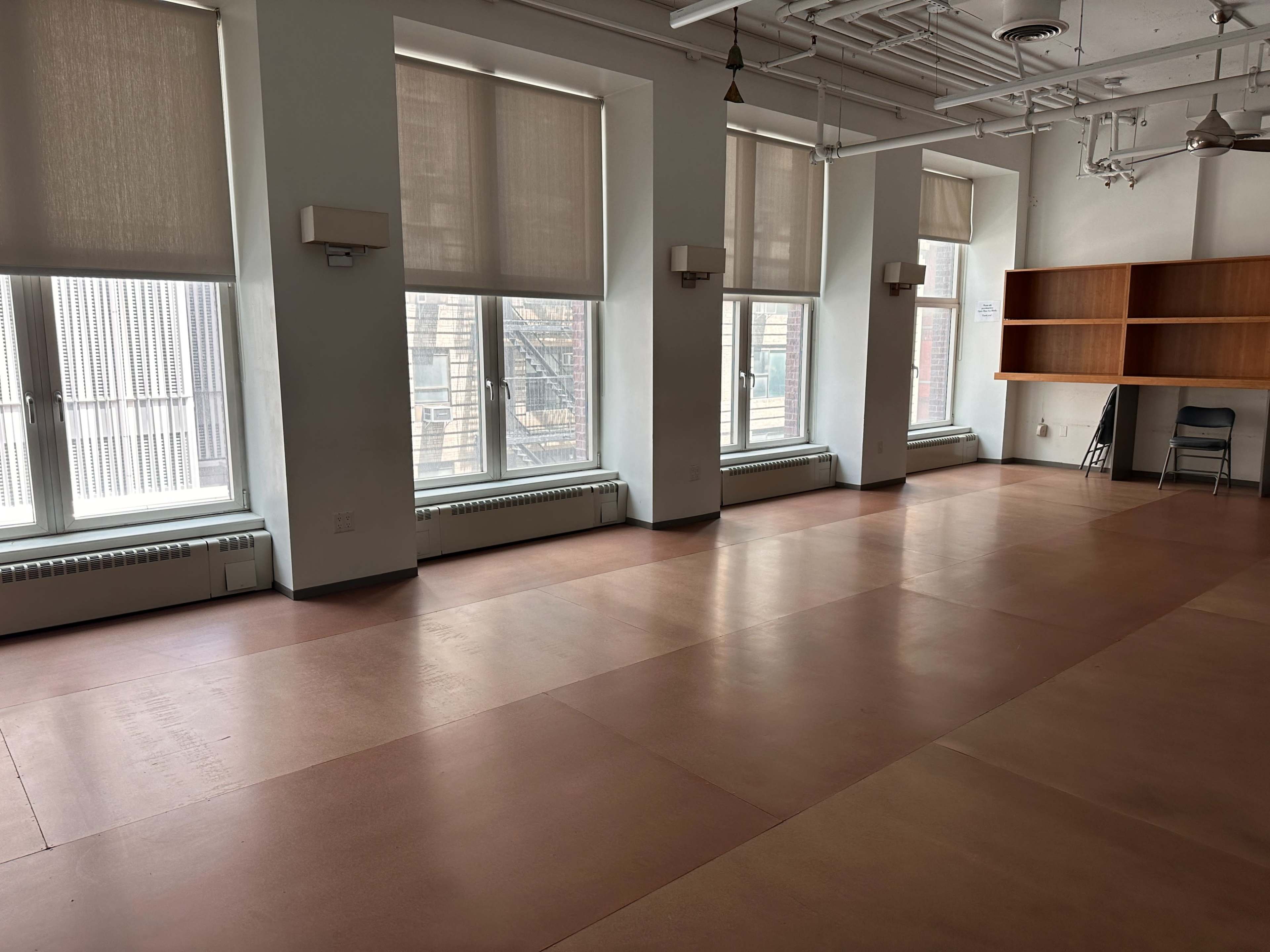 An empty room with large windows, a light-colored floor, and a wooden shelf against one wall.