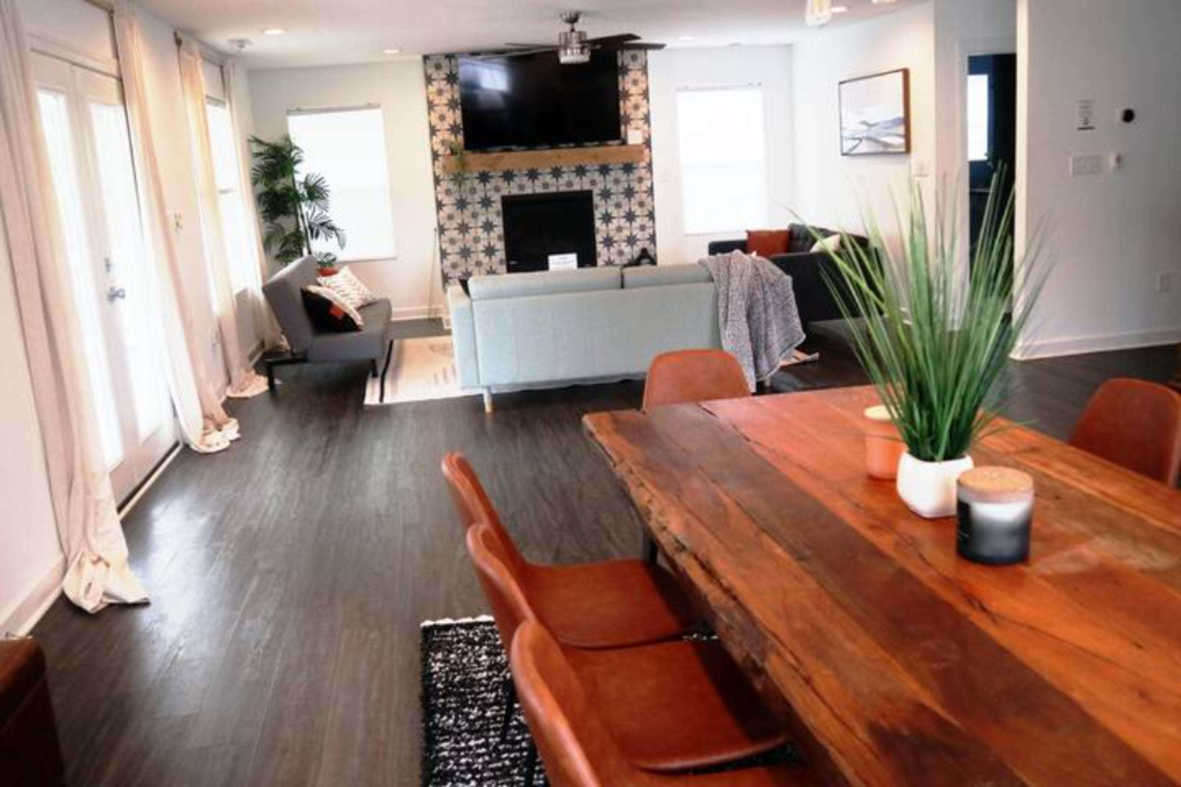 The image shows a modern living and dining area featuring a wooden dining table, comfortable seating arrangements, and a fireplace with a patterned backdrop.