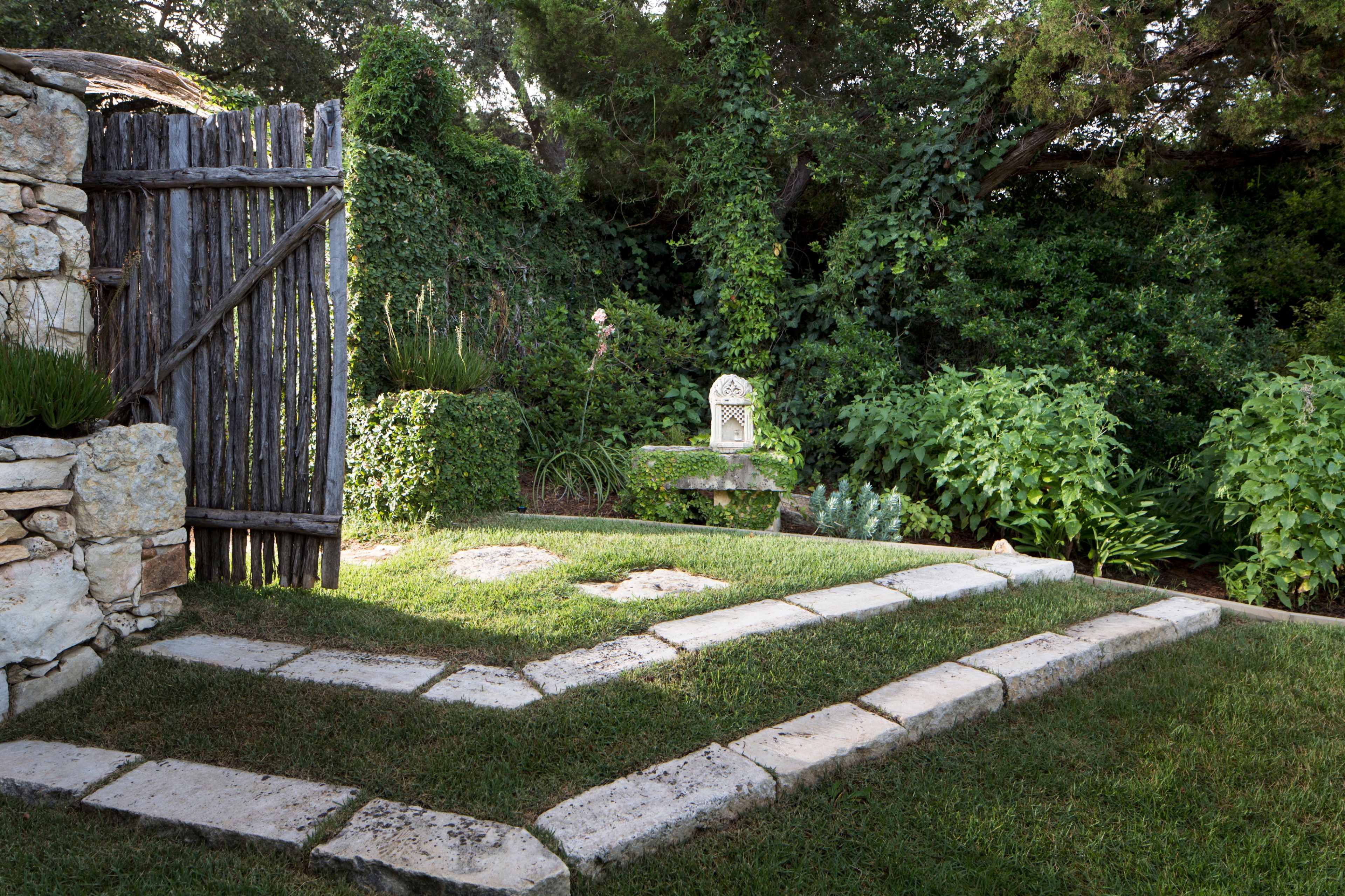 A stone pathway leads through a garden, bordered by lush greenery and a wooden fence.