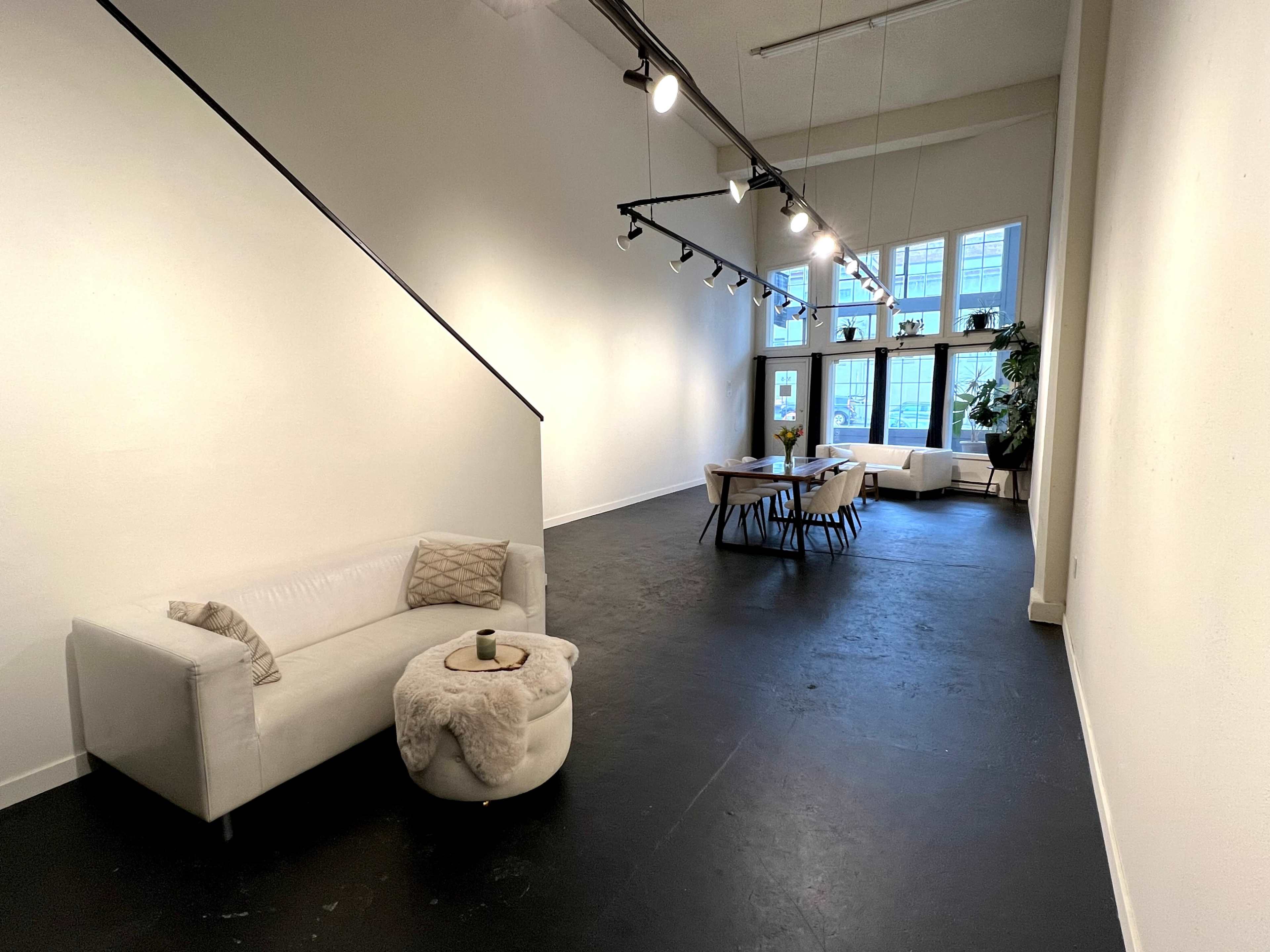 A modern, open interior space featuring a white sofa, a circular ottoman, a large dining table, and large windows allowing natural light.