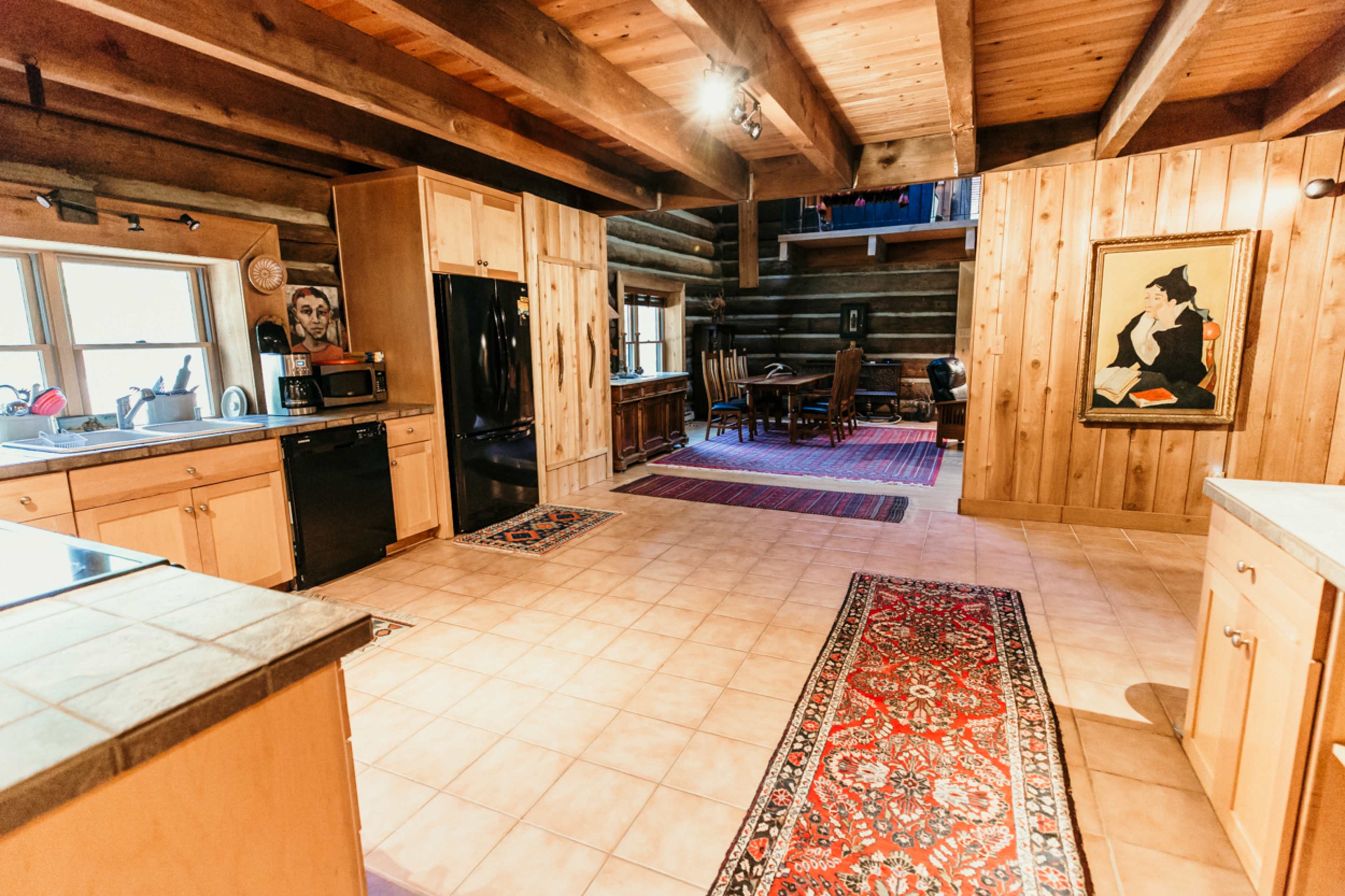 A spacious kitchen with wooden beams and log walls, featuring a tiled floor, modern appliances, and a dining area visible in the background.