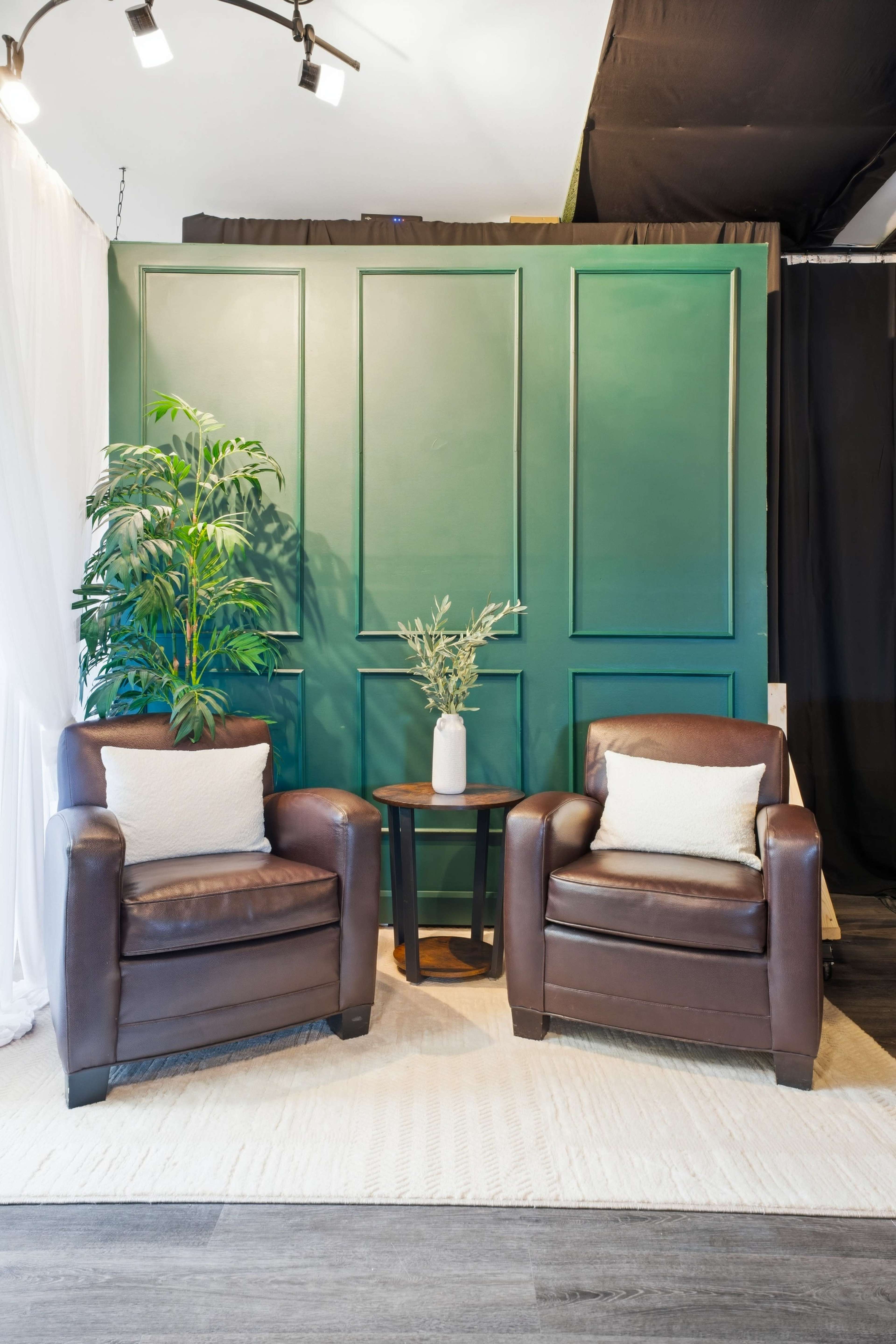 The image features a cozy seating area with two brown leather chairs, a small round table, and a green paneled backdrop, accompanied by a potted plant and a decorative vase.