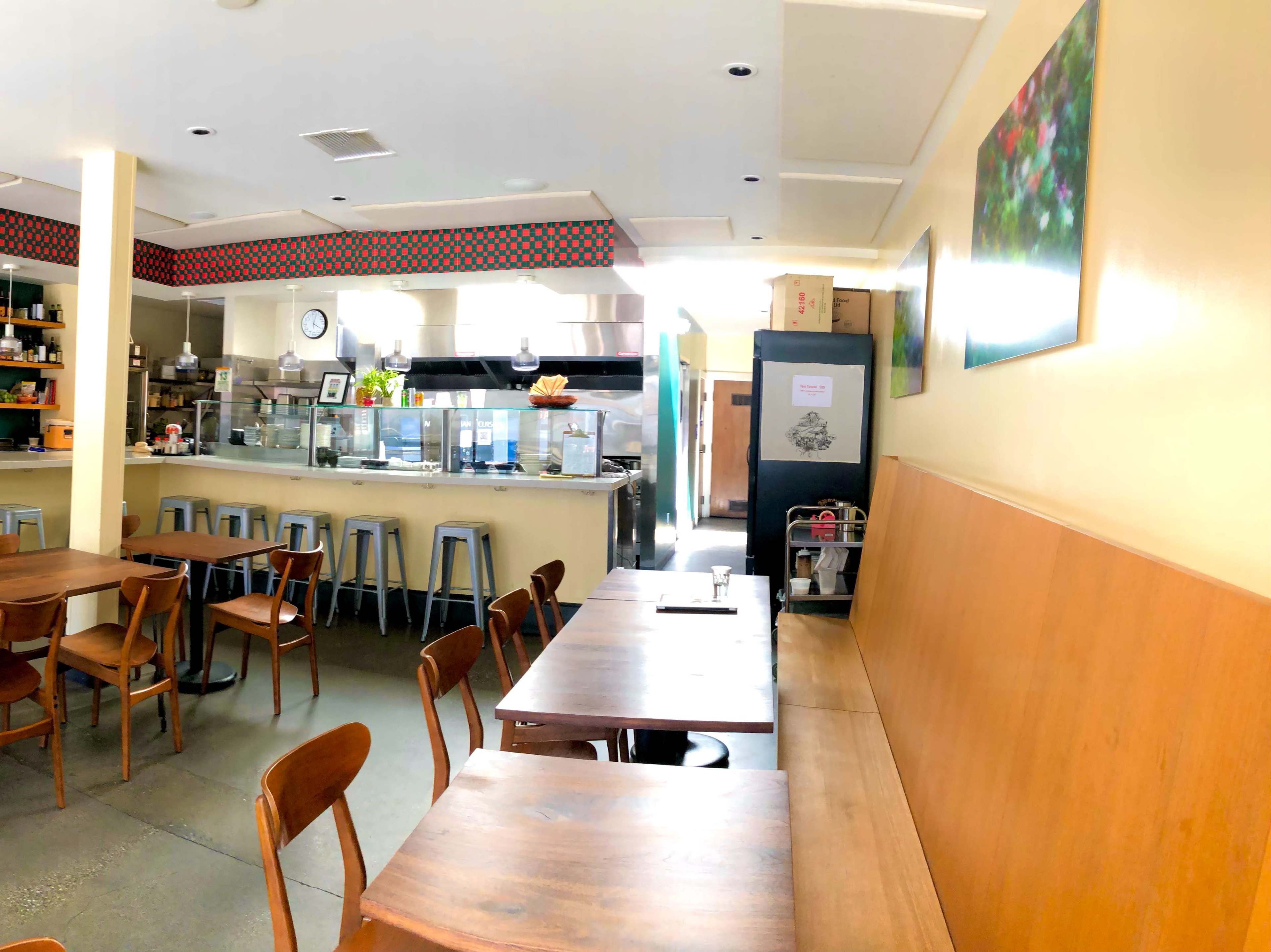 A minimalist cafe interior with wooden tables and chairs, a counter with kitchen equipment in the background, and bright natural light coming through a window.
