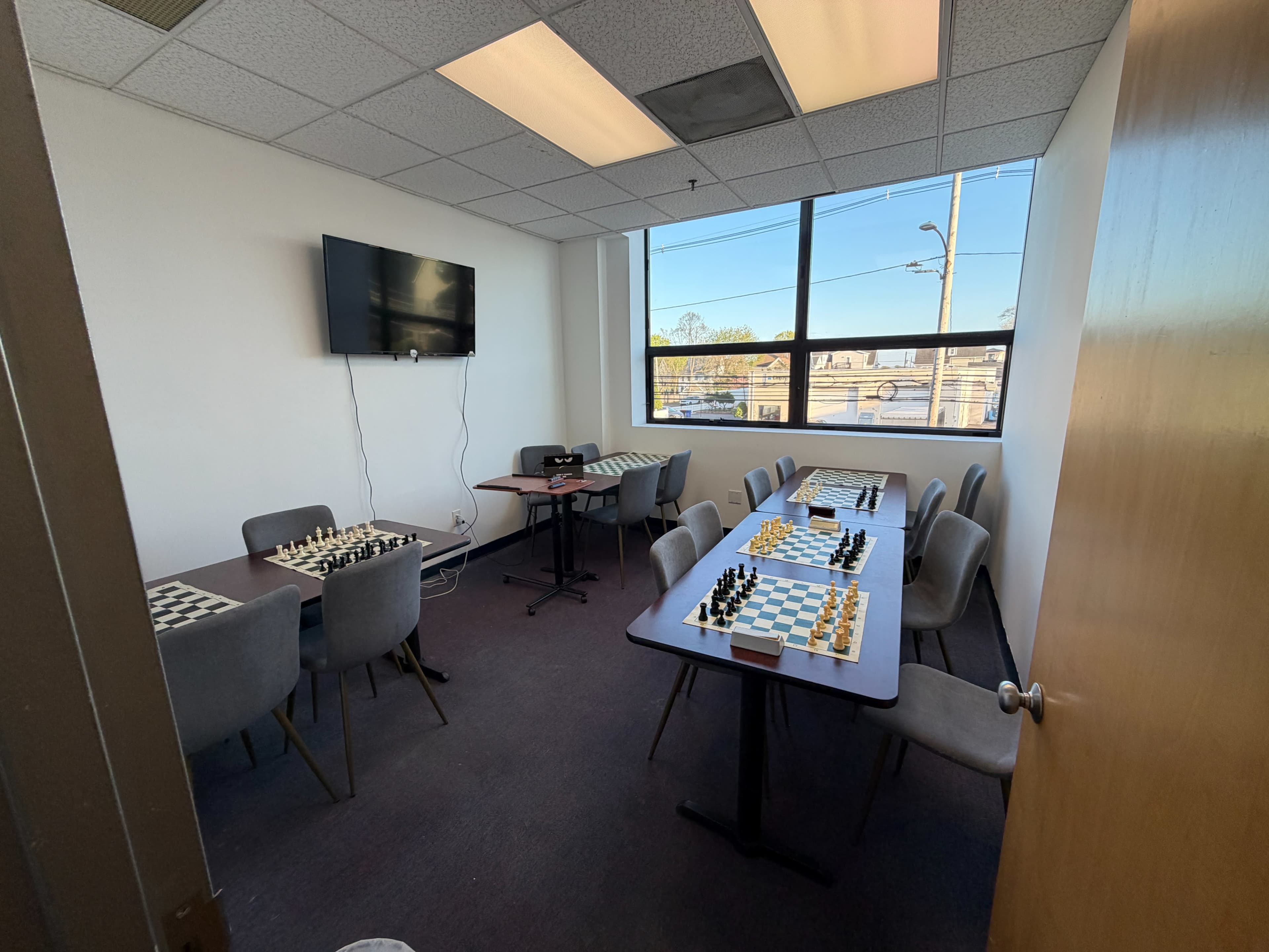The image shows a small meeting room with two tables set up for chess, accompanied by chairs and a wall-mounted television.