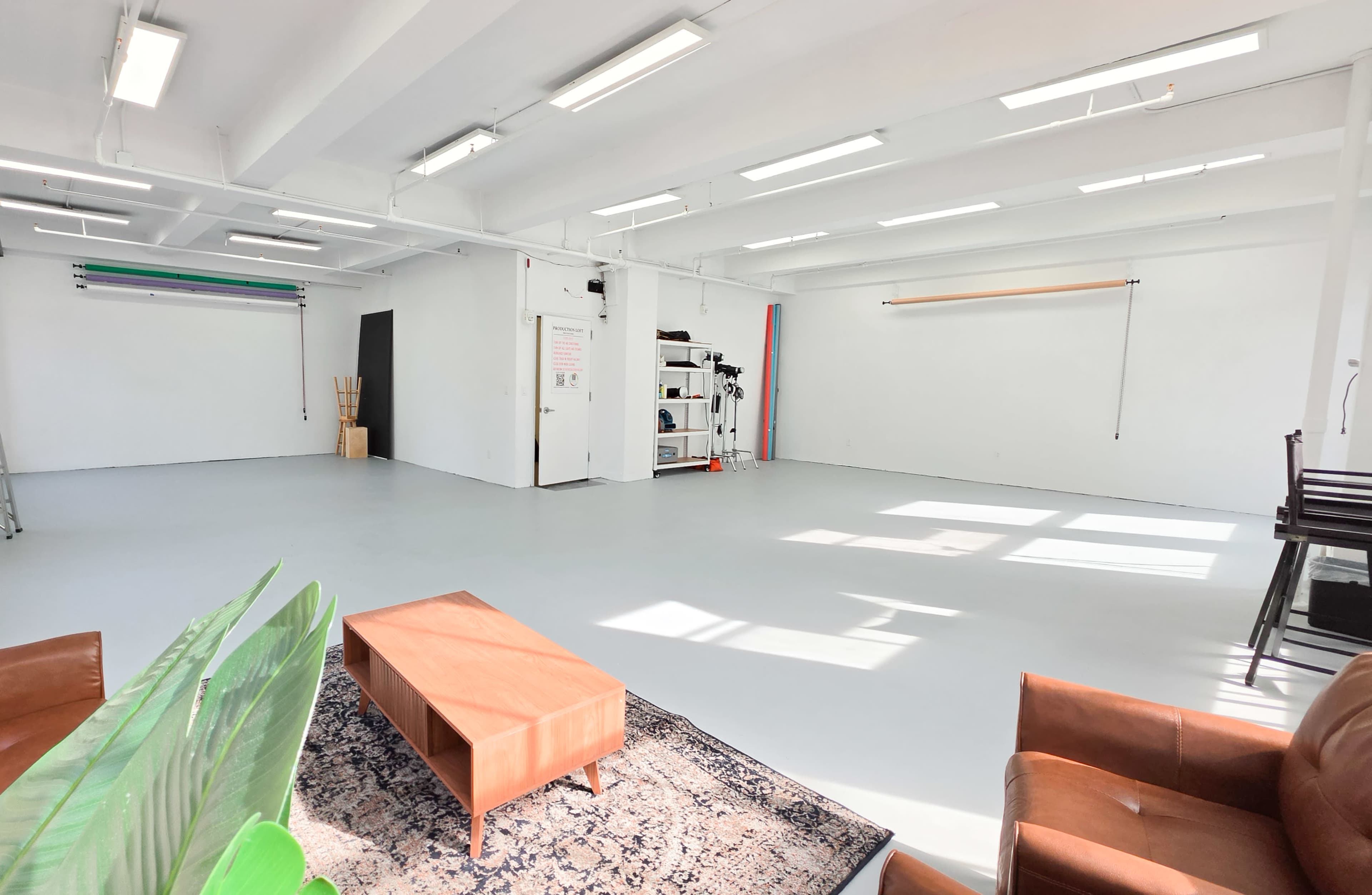 The image shows a spacious, well-lit studio with white walls, a polished floor, and minimal furniture including a brown leather sofa and a wooden coffee table.