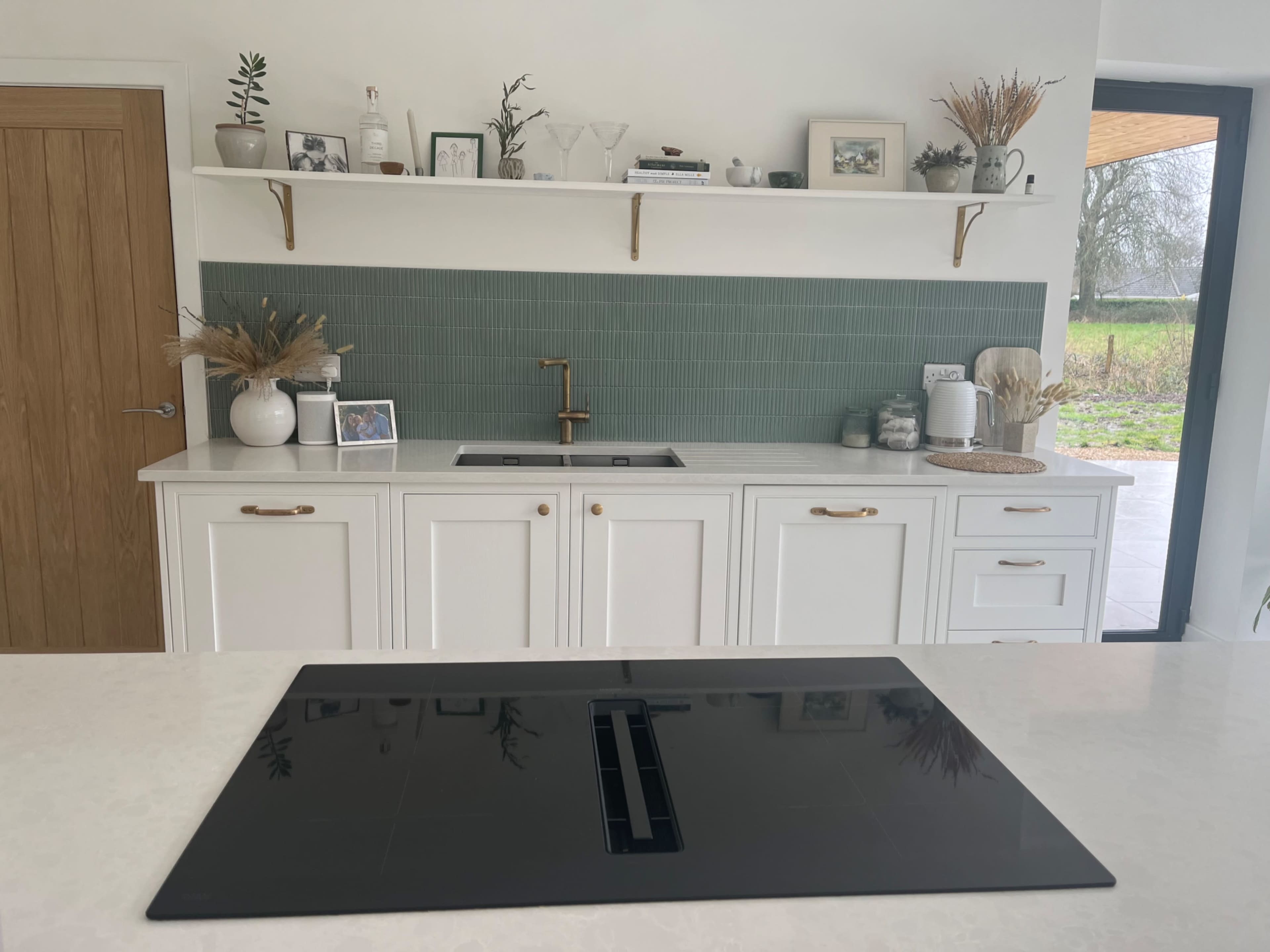 The image shows a modern kitchen with white cabinets, a black cooktop on a white countertop, and a green tiled backsplash decorated with plants and kitchenware on floating shelves.