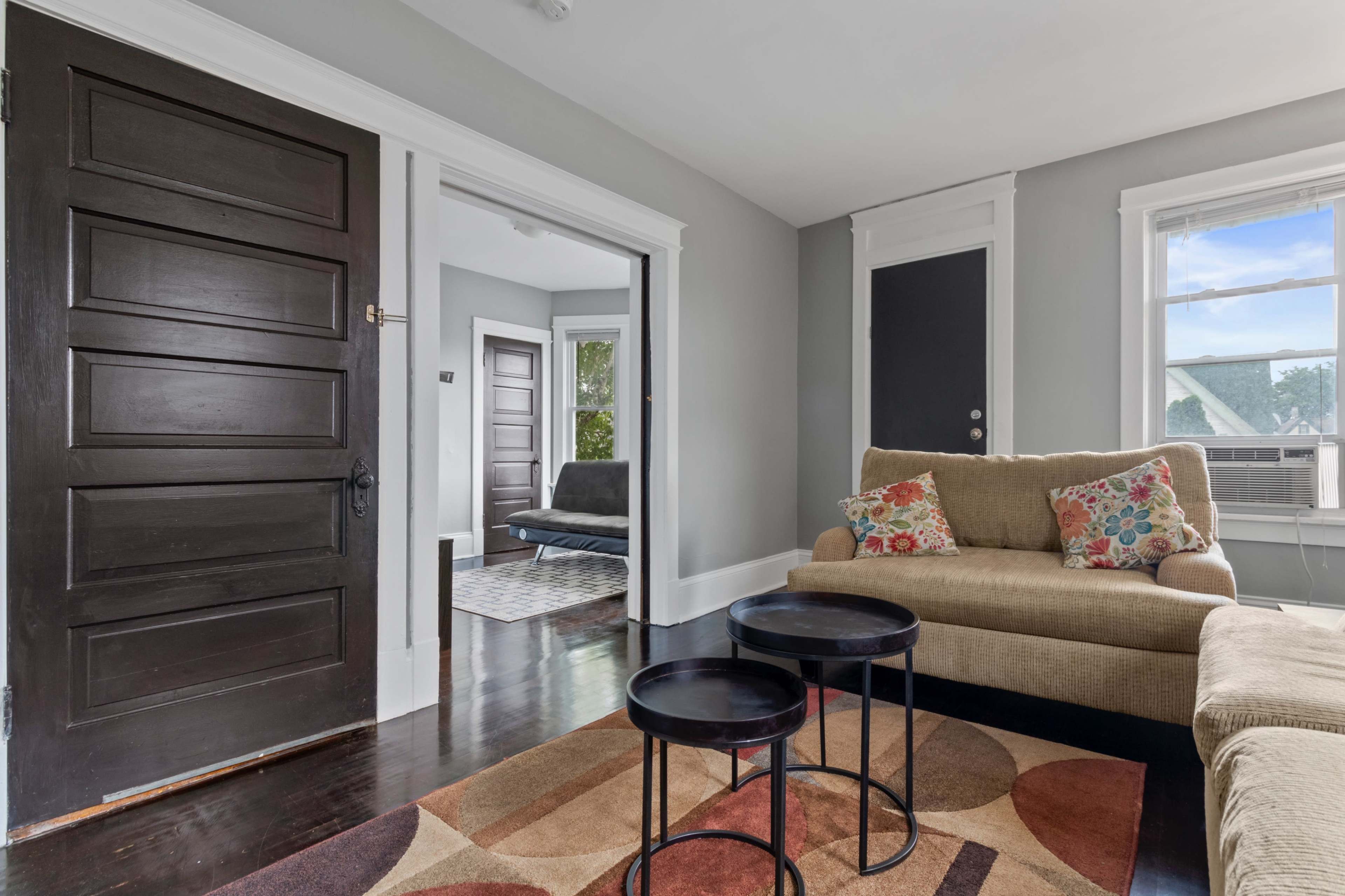 A living room with a beige couch, a rug with circular patterns, two small black tables, and a doorway leading to another room.