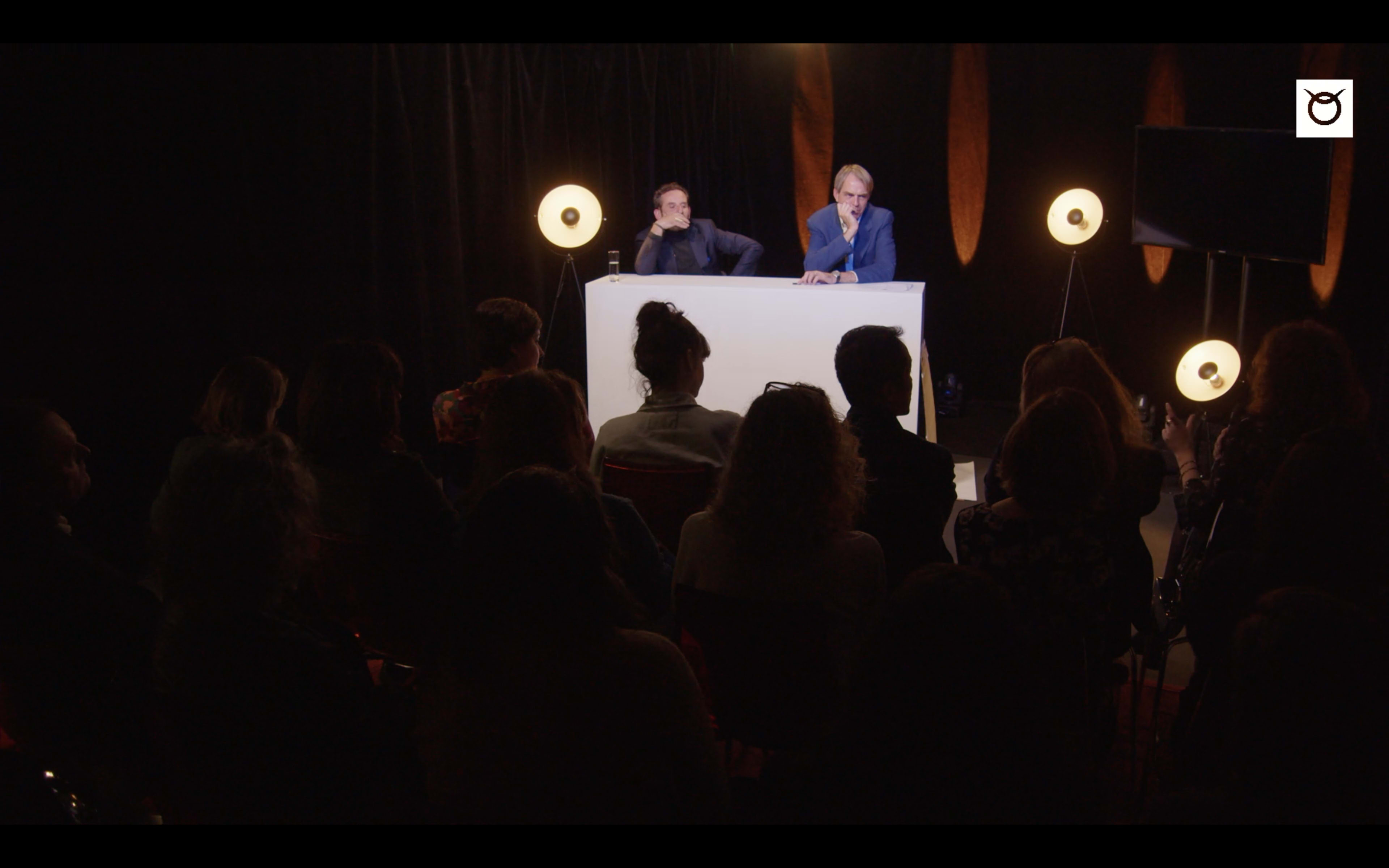 Two speakers sit behind a table in front of an audience, with a dark backdrop and lights positioned around the stage.