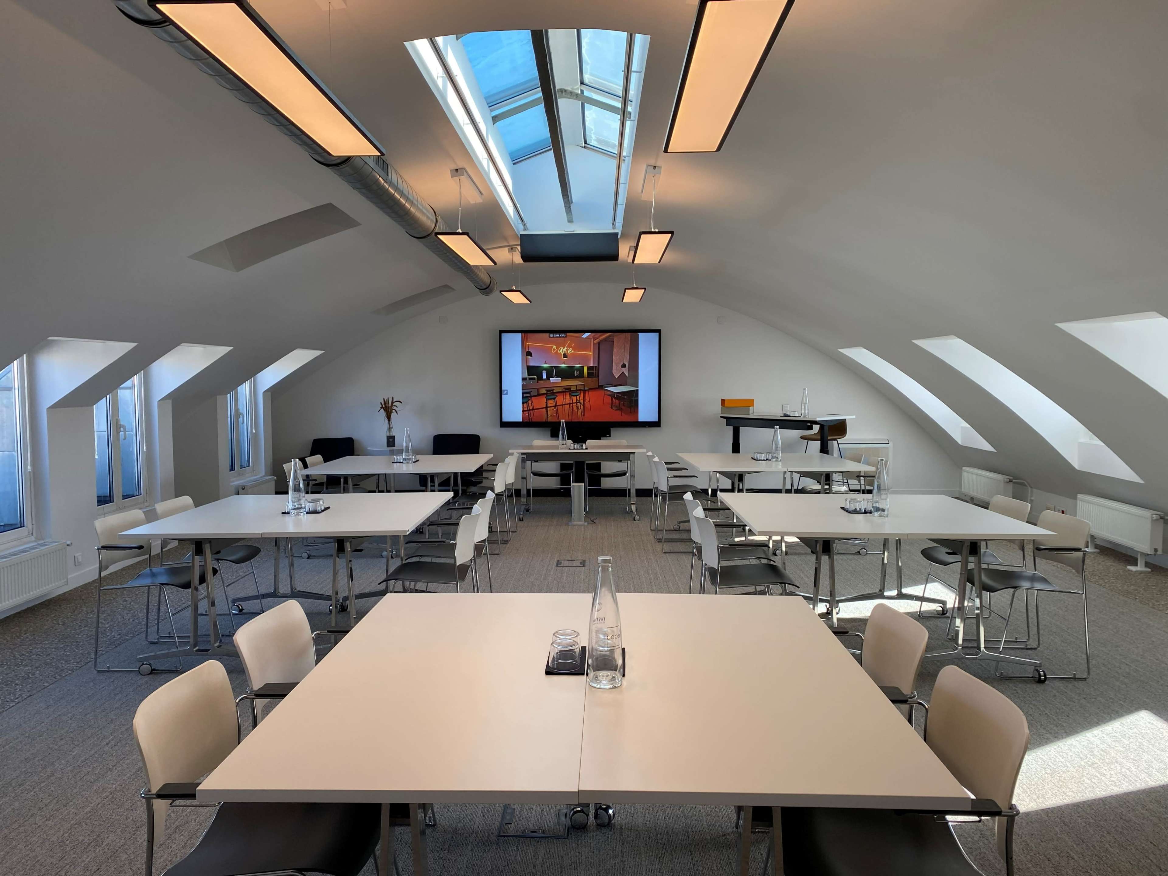 A bright, modern meeting room features several white tables and chairs arranged for a presentation, with large windows and a screen displaying a colorful image on the wall.
