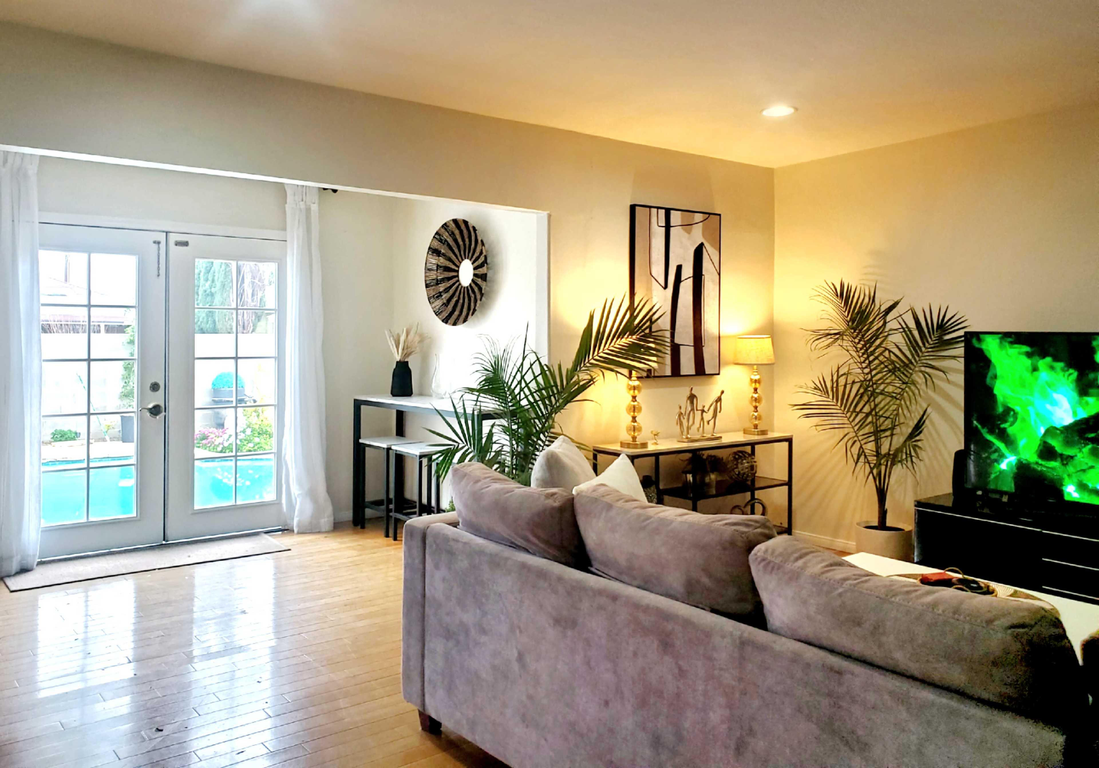 A living room features a gray sofa, a television, a table, and plants, with large windows opening to a pool area.