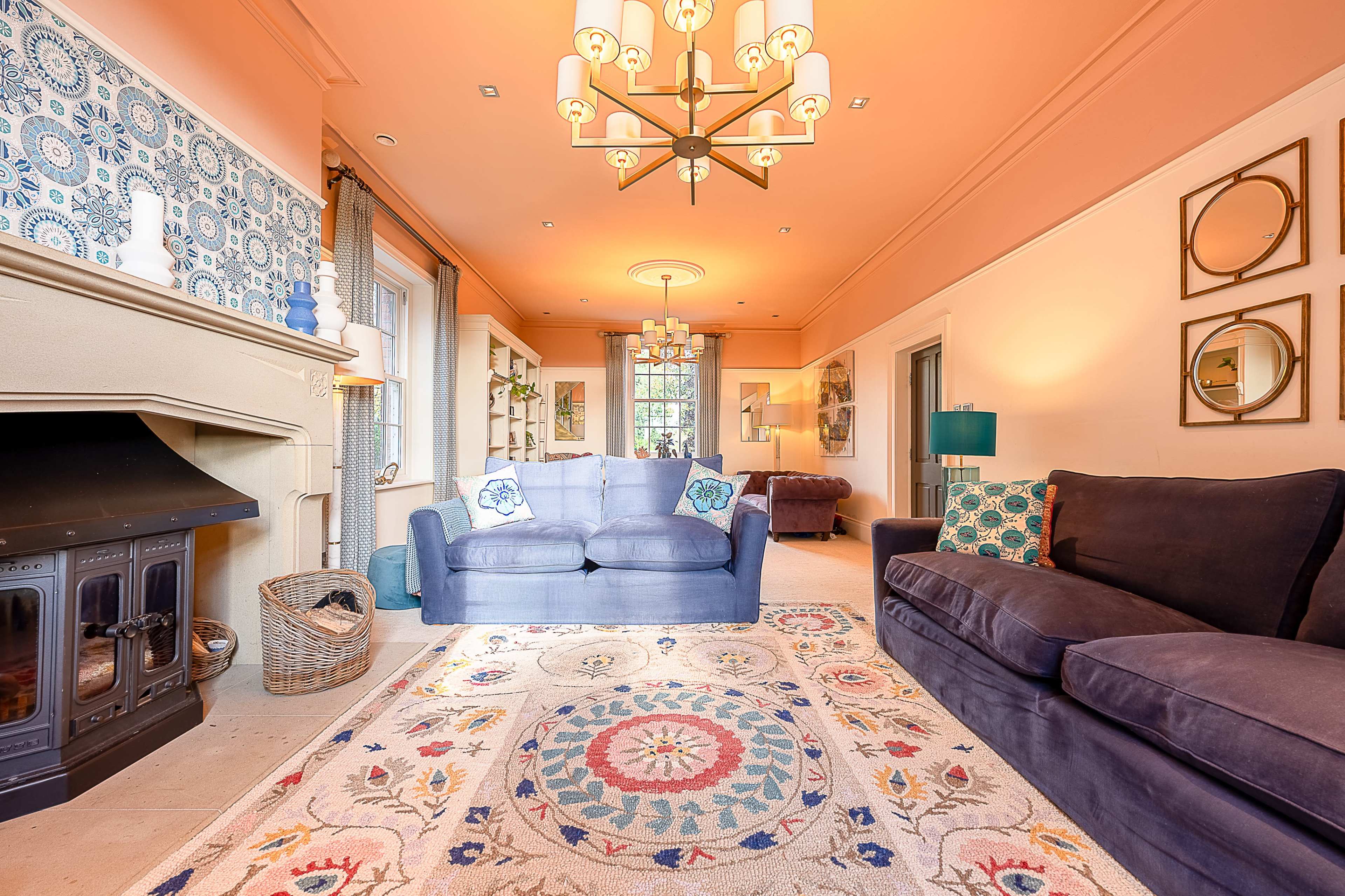 The image shows a living room featuring two sofas, a patterned area rug, a fireplace, and decorative wall elements, all under a chandelier.