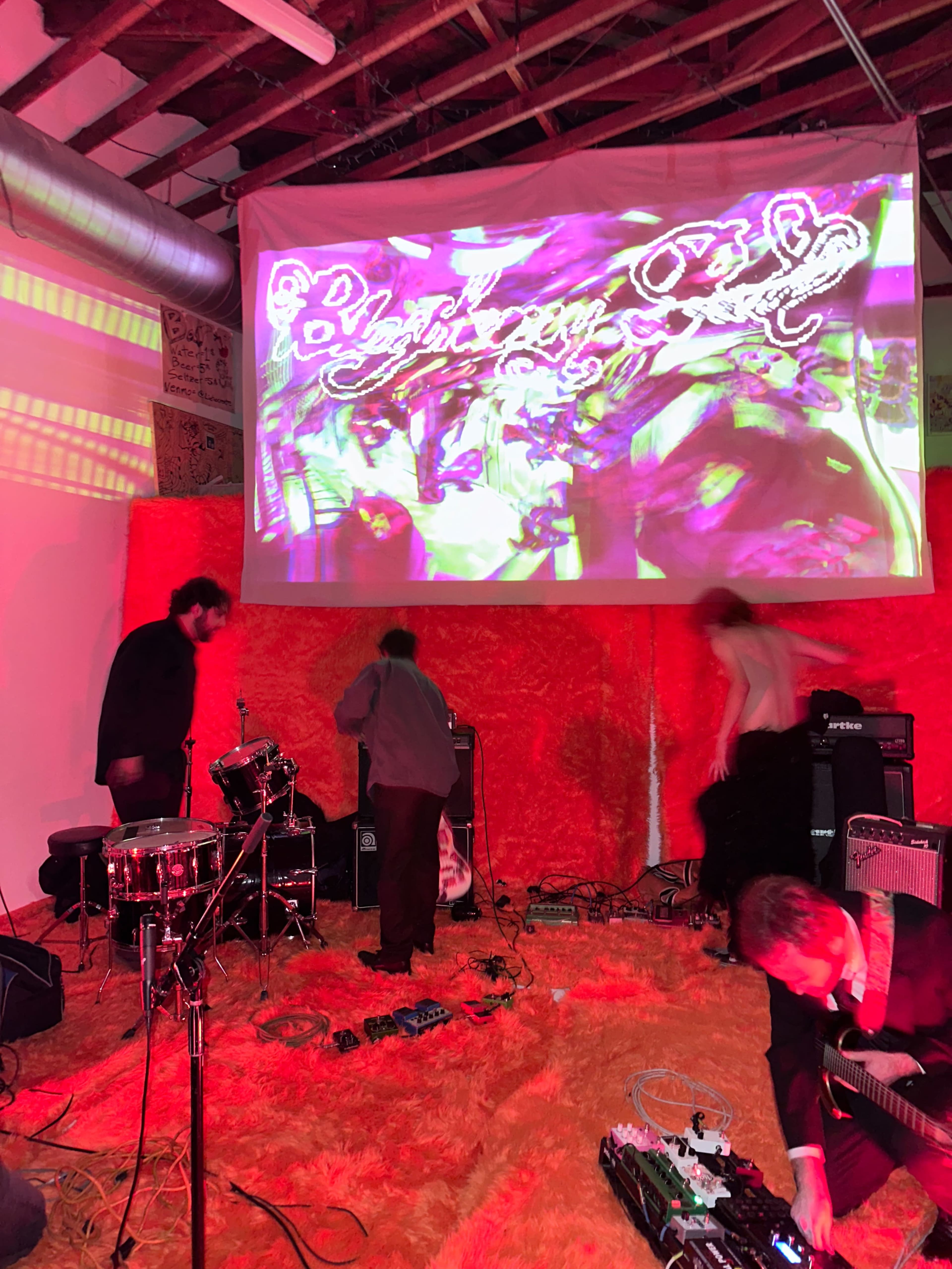 A group of musicians prepares for a performance in a dimly lit space with orange decor and a large projected visual display.