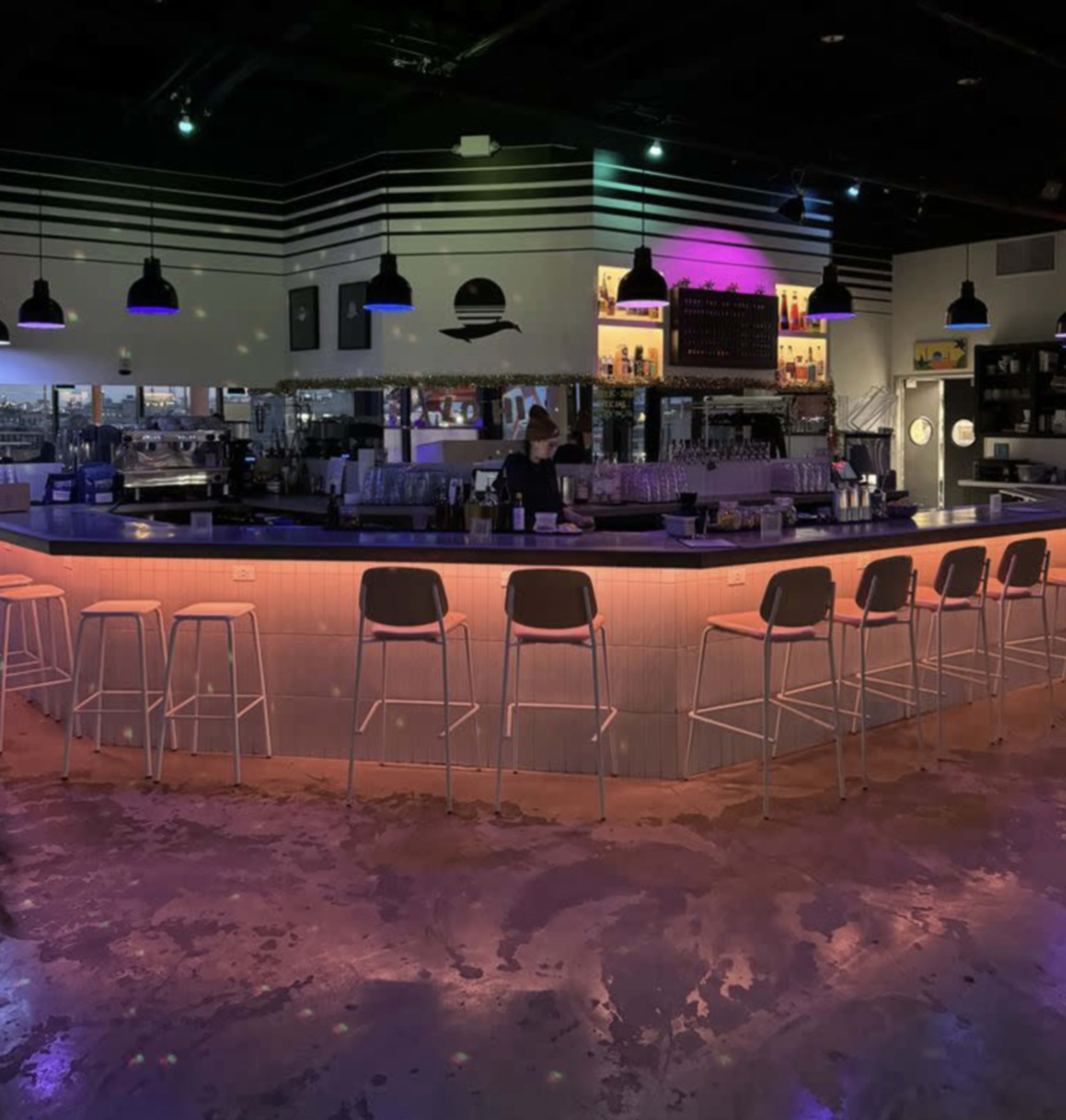 A modern bar features a curved countertop with illuminated edges, surrounded by high stools and colorful lighting.