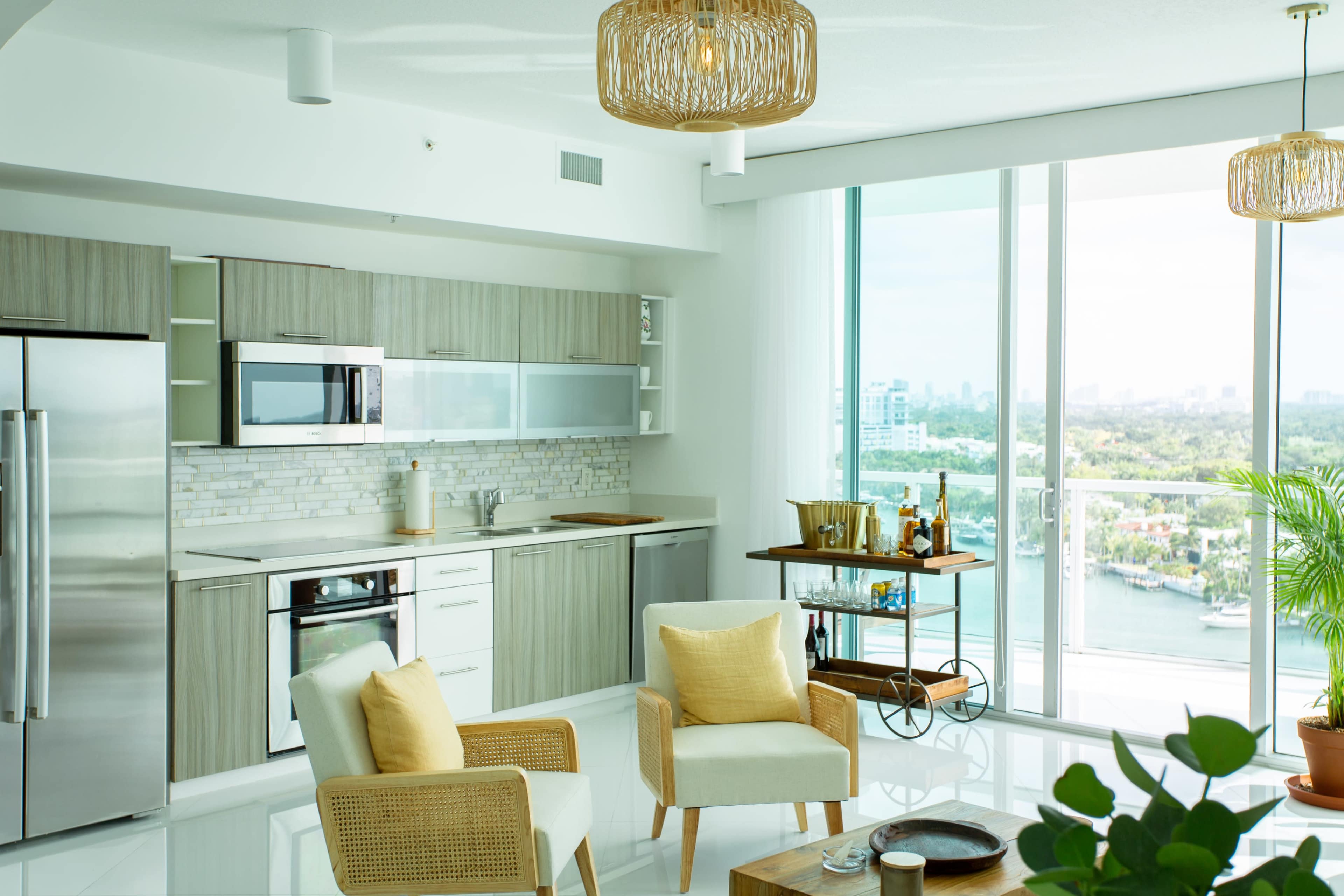 A modern kitchen and living area feature light wood cabinetry, stainless steel appliances, and large windows overlooking a cityscape.