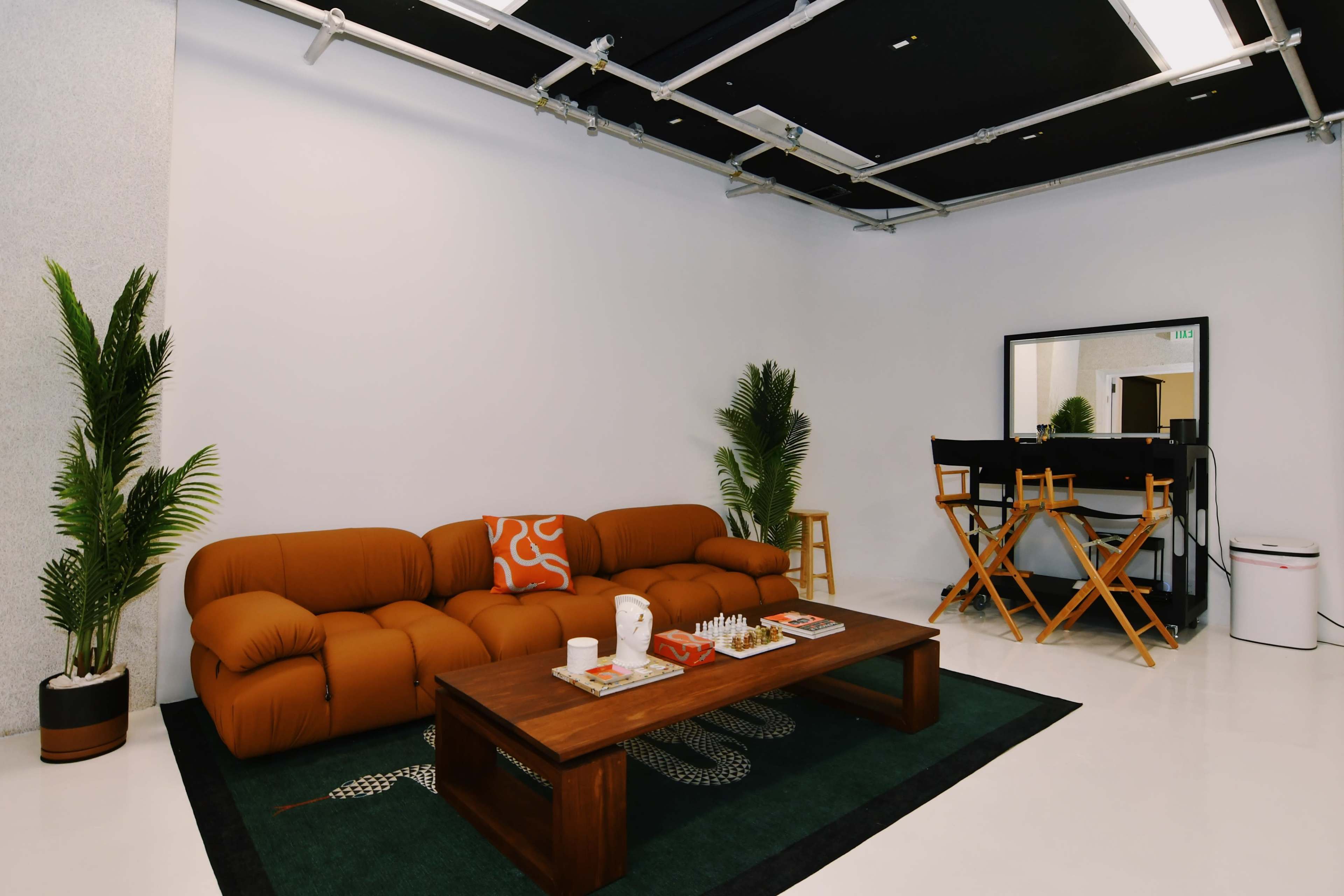 A modern living room with a brown sectional sofa, a wooden coffee table, and a makeup station with director's chairs against a white wall.