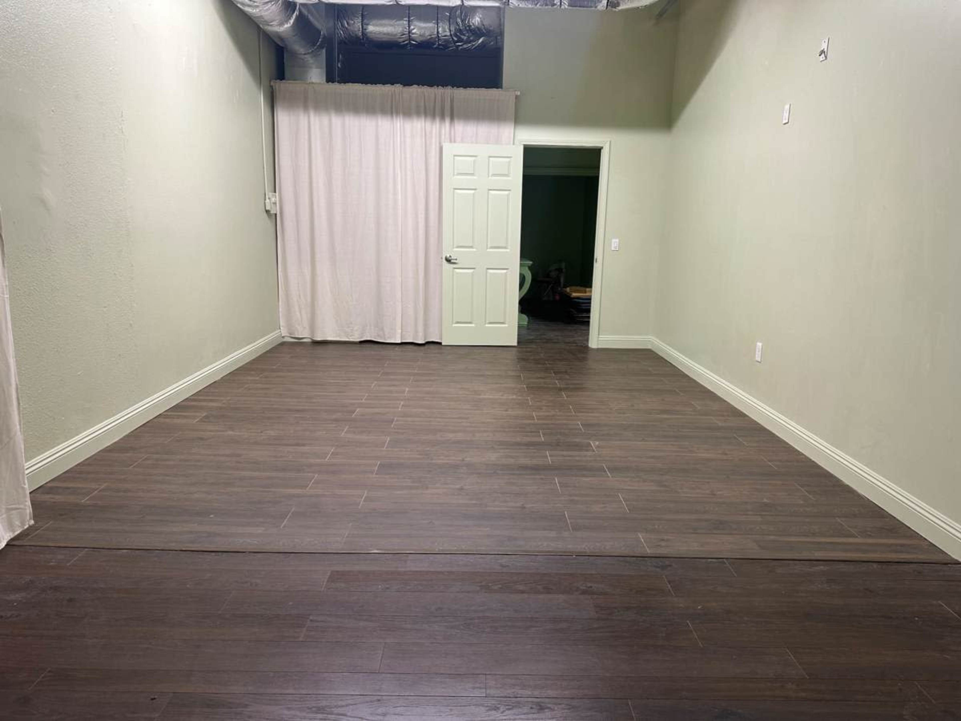 The image shows a spacious, empty room with light green walls, a brown wooden floor, and an open door leading to a darkened area.