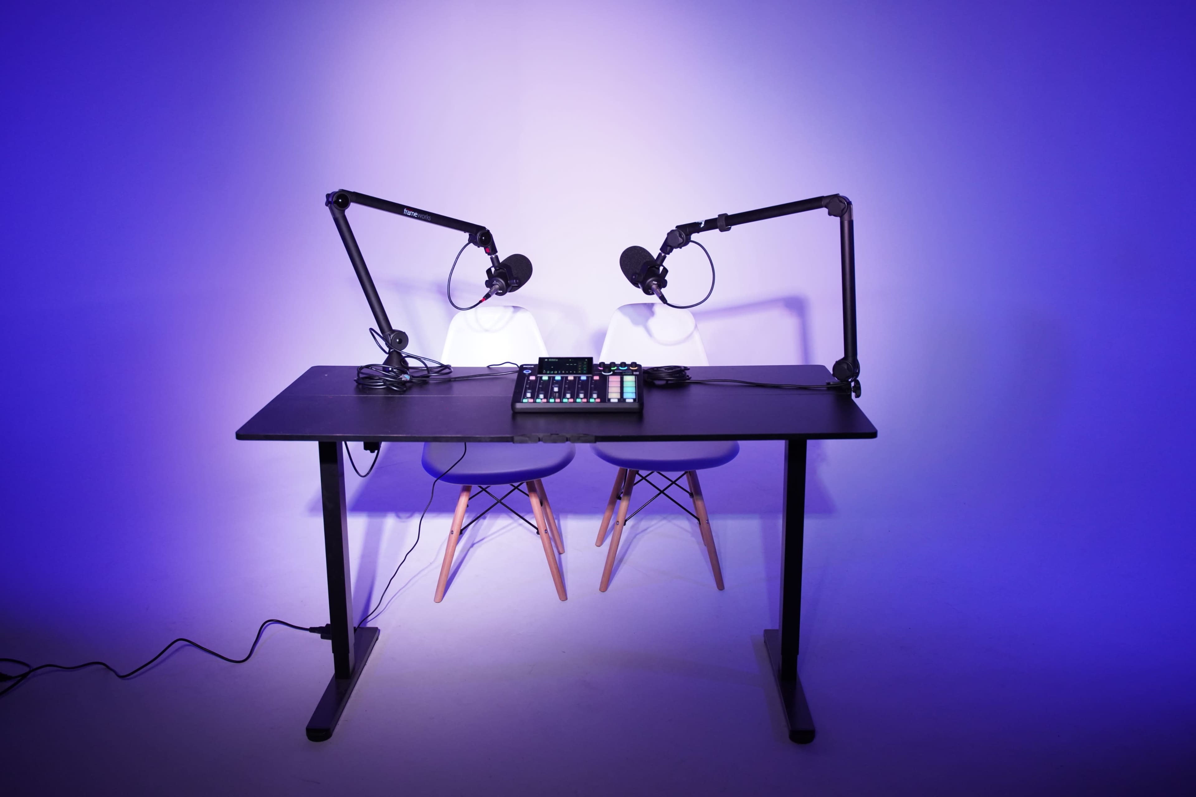 A podcasting setup featuring two microphones on adjustable arms, a control panel, and two chairs on either side of a black table, all against a purple backdrop.