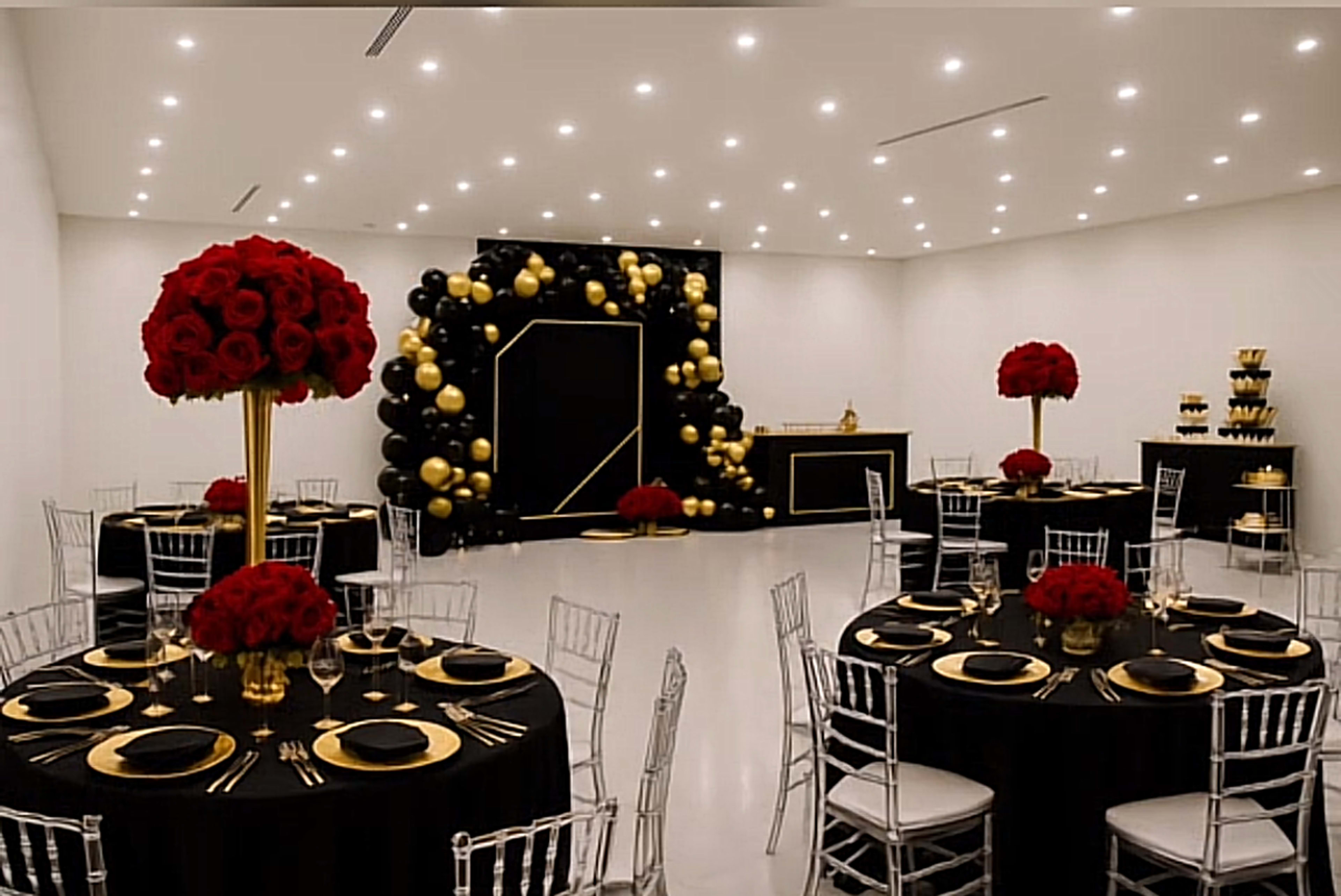 A banquet hall features round tables with black tablecloths and centerpieces of red roses, set against a backdrop of gold and black balloons.