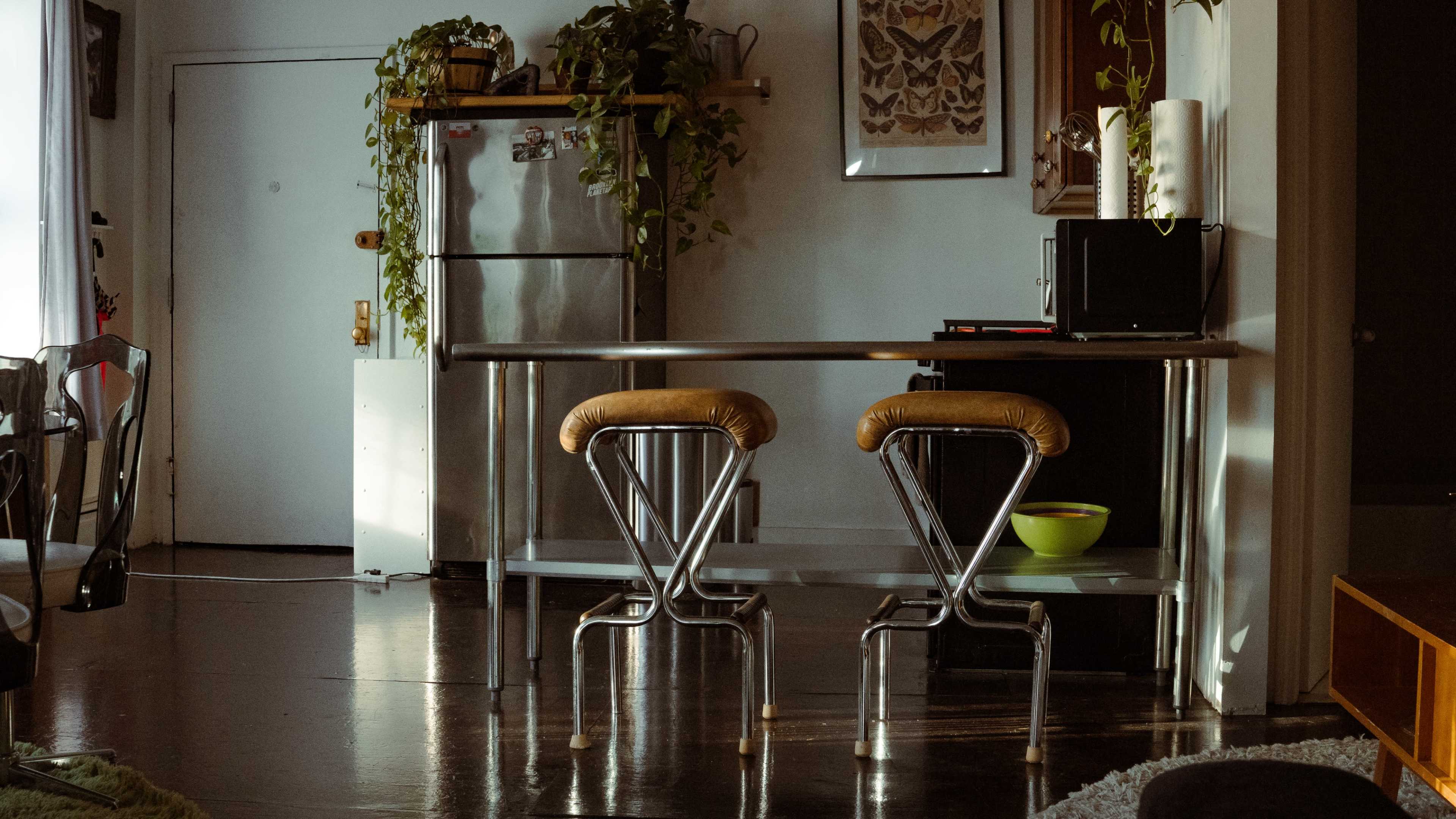 A minimalistic kitchen area features a stainless steel table with two stools and a green bowl, alongside a refrigerator and a microwave.