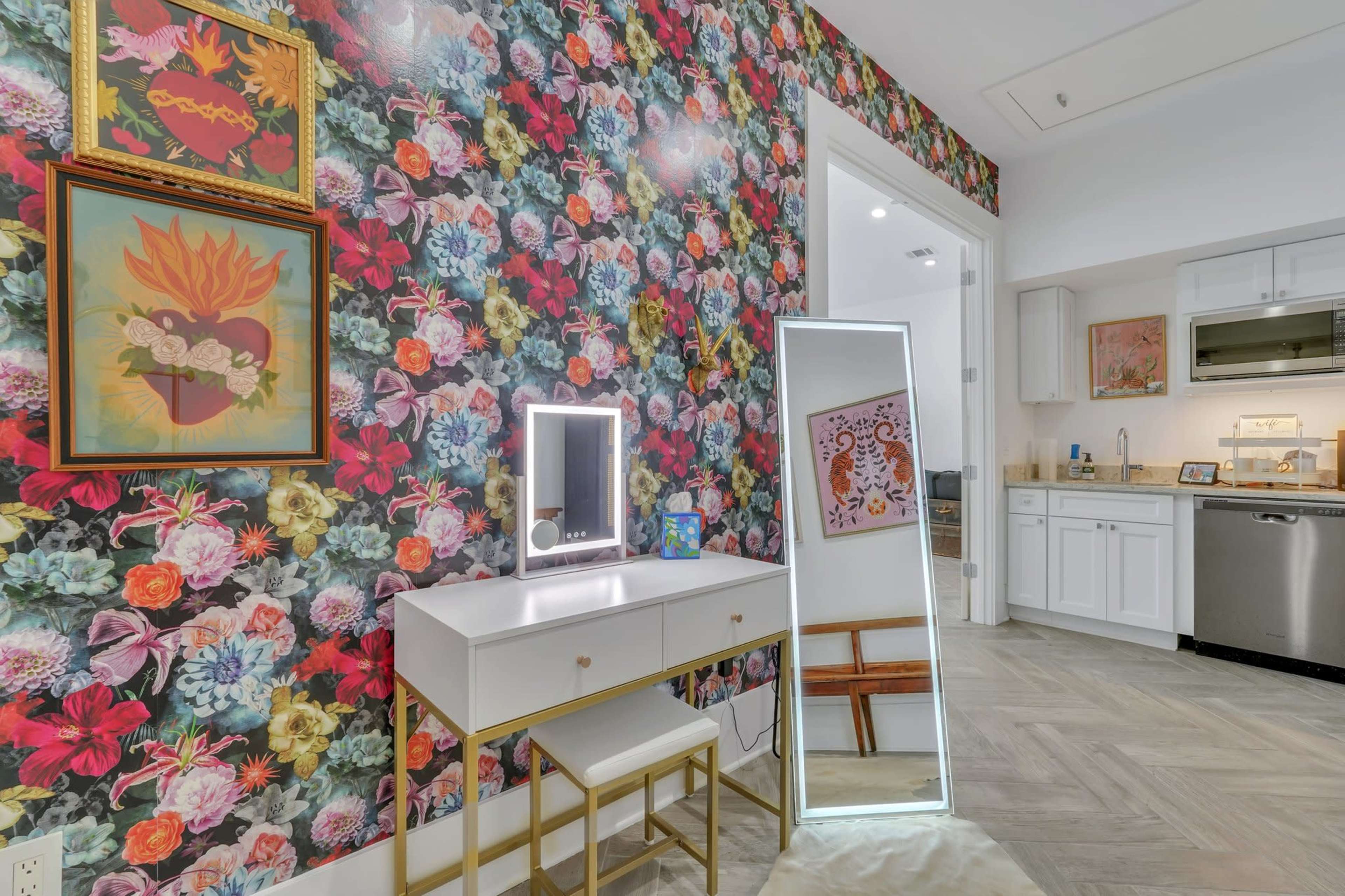 A brightly decorated room featuring a floral wallpaper, a white vanity with a mirror, and a kitchen area in the background.