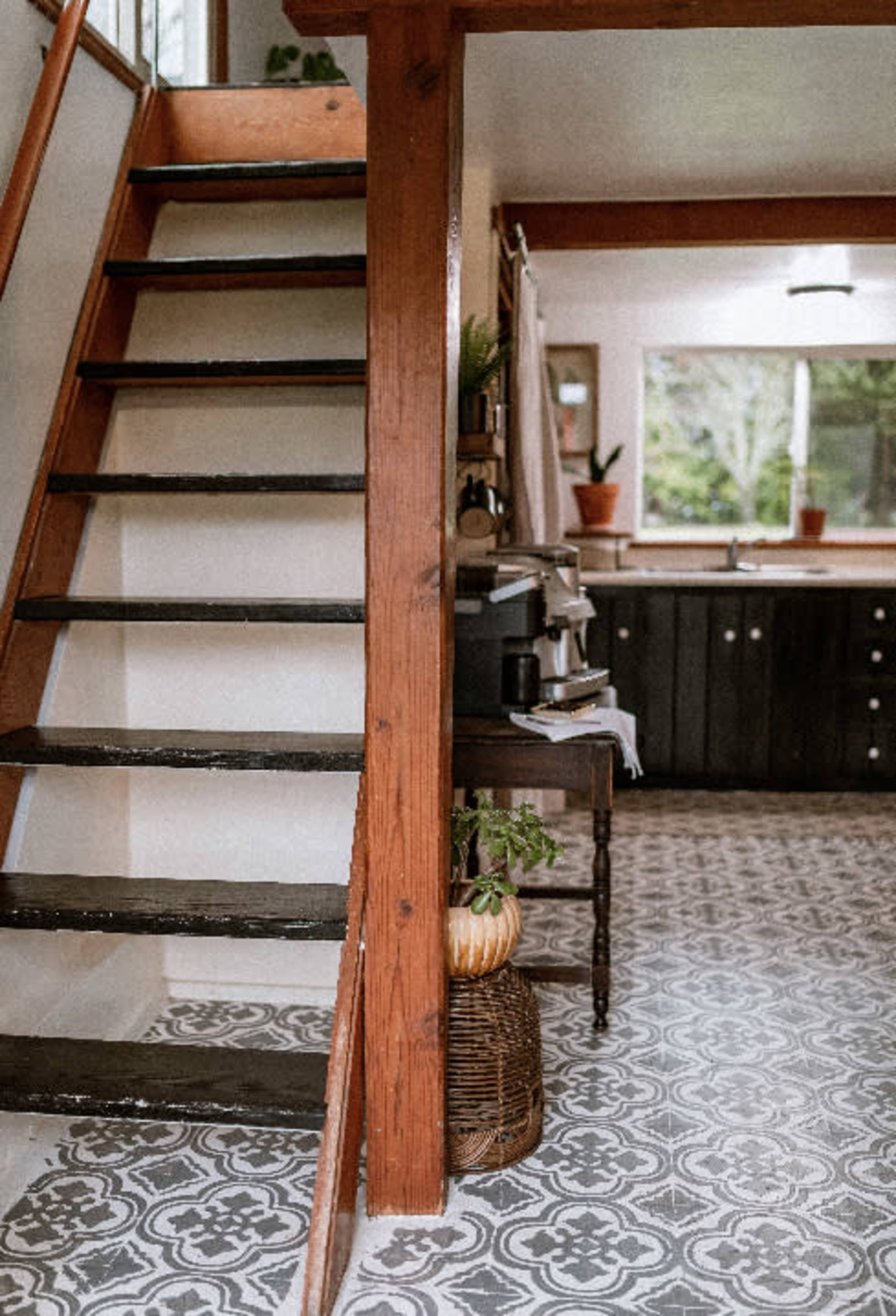 The image shows a kitchen area with a staircase leading to an upper level, featuring black cabinetry, a patterned floor, and potted plants.