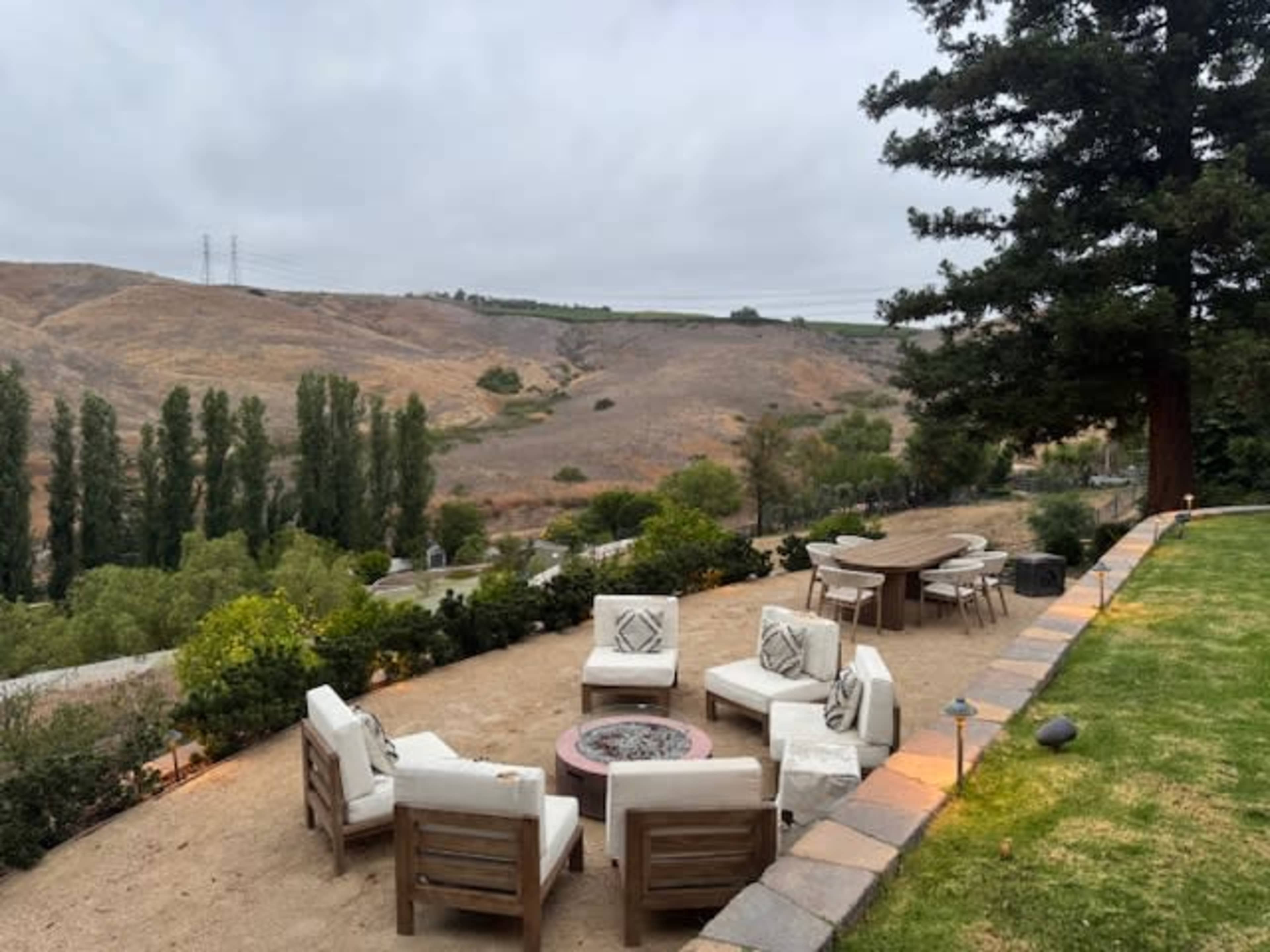 A patio scene features wooden furniture arranged around a fire pit, overlooking a hilly landscape with scattered trees.