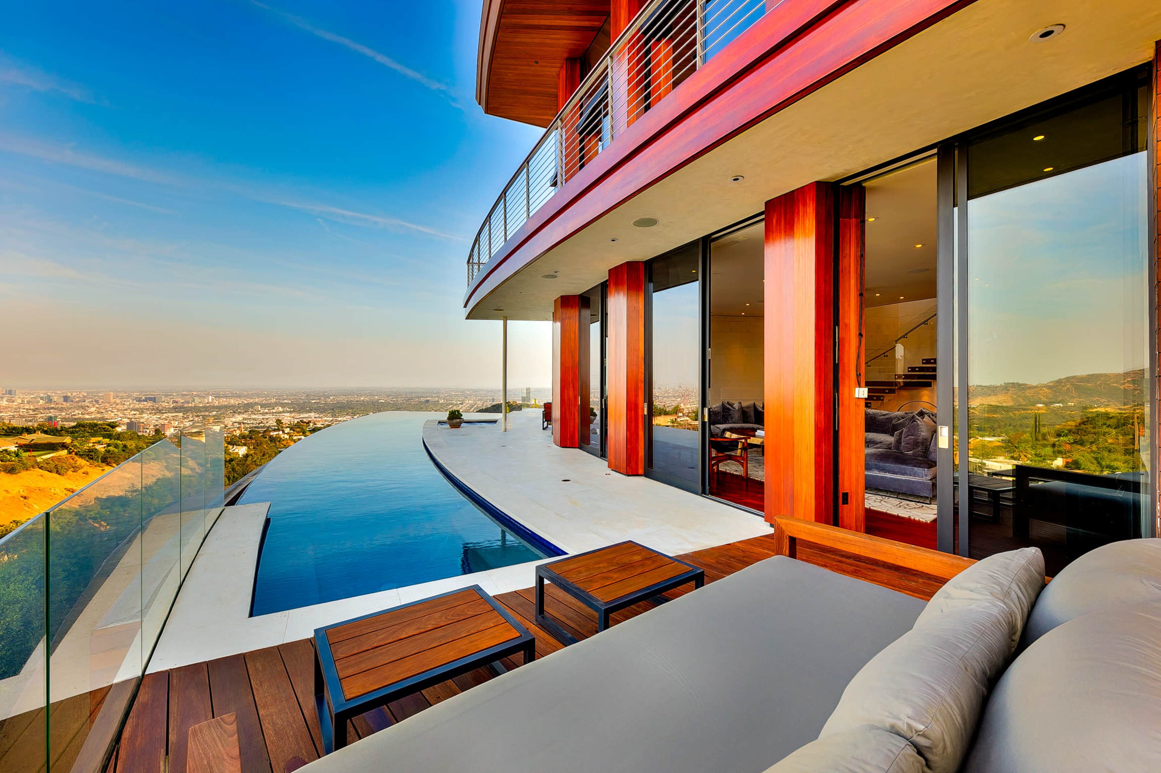 The image shows a modern villa with large glass walls, an infinity pool, and a balcony overlooking a scenic view.