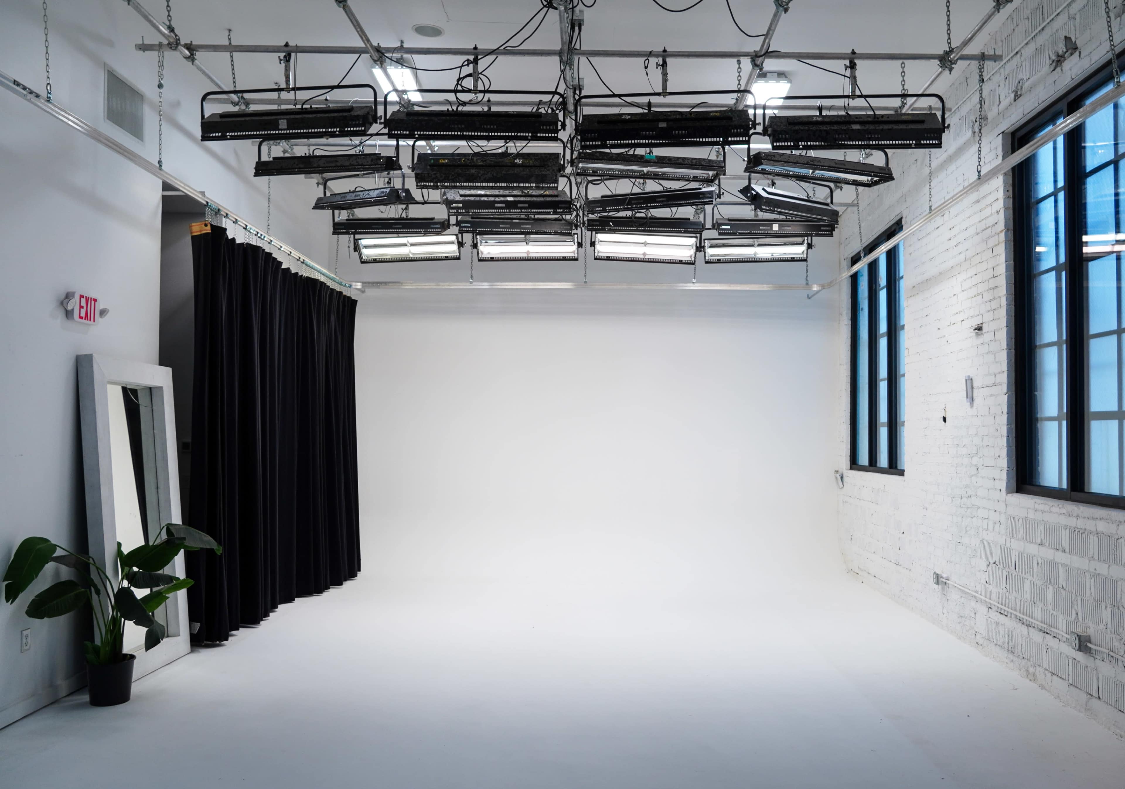 The image shows a spacious, empty studio with a white wall, hanging lights from the ceiling, and a large window alongside a black curtain and a potted plant.